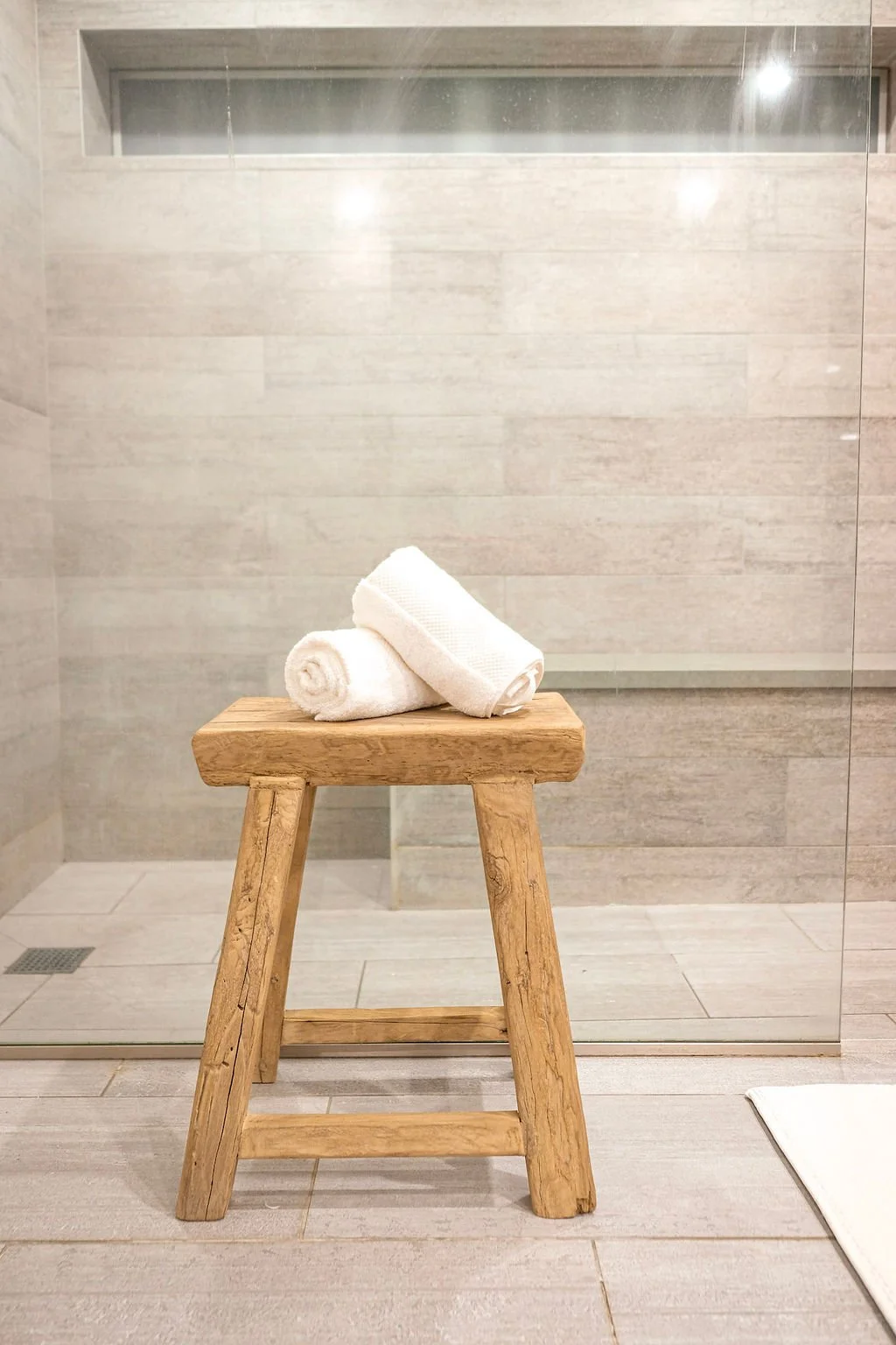 A wooden stool with two rolled white towels placed on it in front of a glass shower enclosure with beige tile walls and a small window.