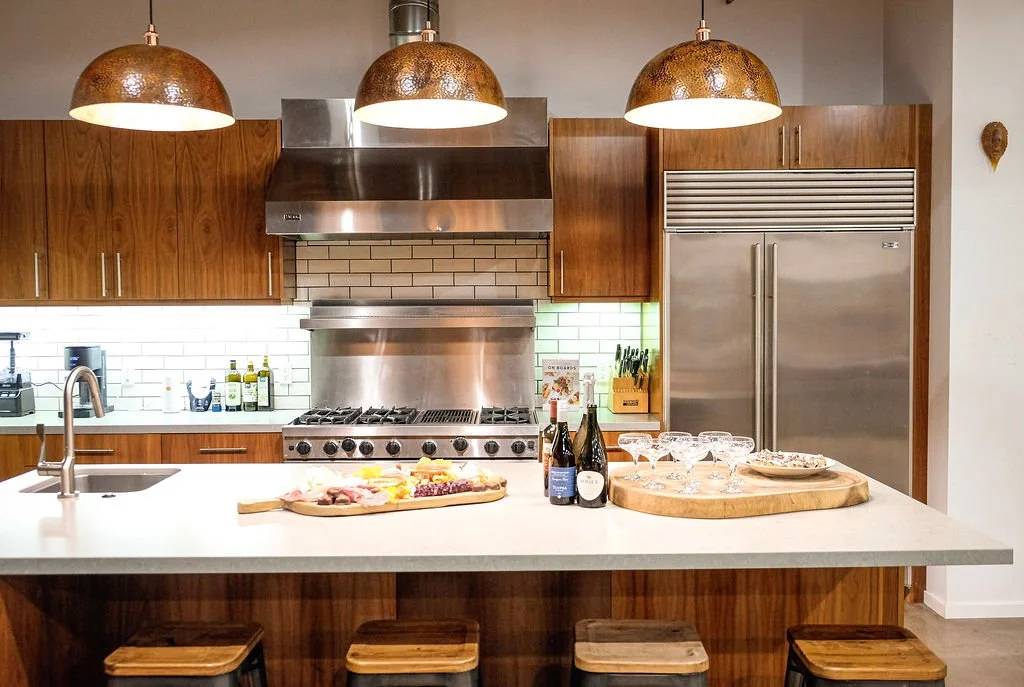 Modern kitchen with wooden cabinets, stainless steel appliances, and a white countertop island. On the island, there are bottles of wine and glasses, a wooden serving board with food, and a plate. Overhead are three copper-colored pendant lights.