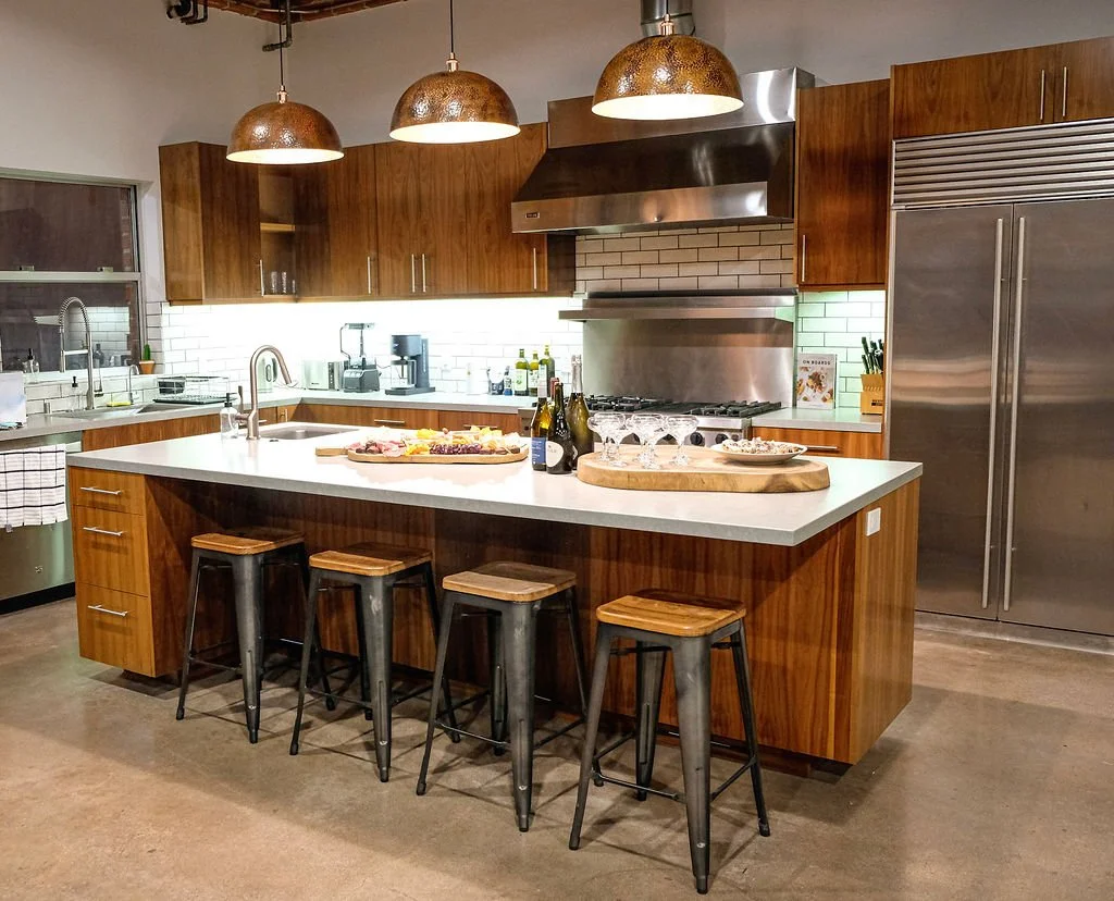 Modern kitchen with wooden cabinets, stainless steel appliances, a white island countertop with four metal and wood stools, pendant lights, and a white subway tile backsplash.