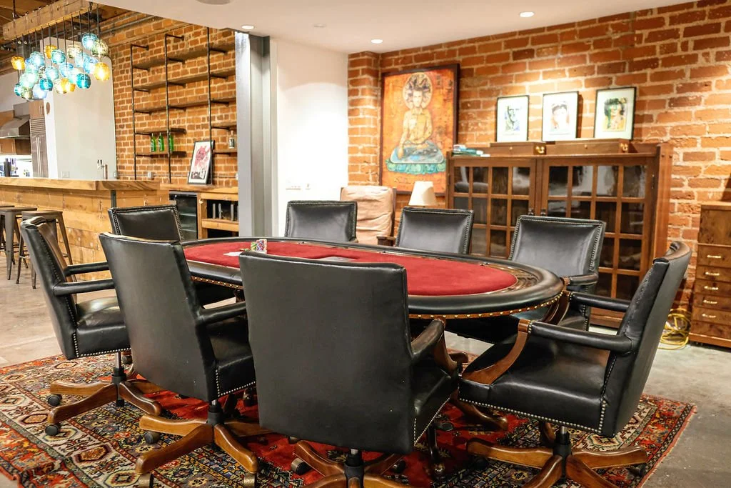 A poker table with eight black leather chairs in a room with brick walls, wooden furniture, and colorful artwork.