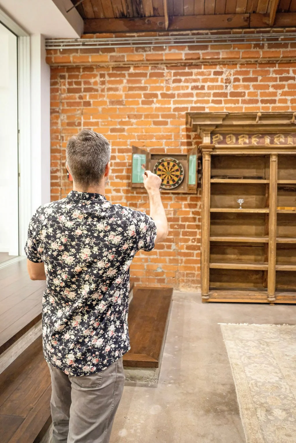 A man with short hair wearing a floral shirt and gray pants is playing darts indoors against an exposed brick wall.