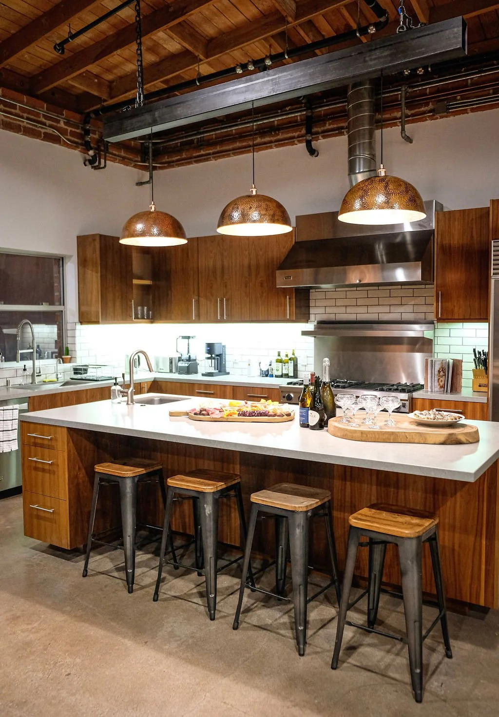 Modern kitchen with a large white island countertop, four metal stools with wooden seats, wooden cabinets, pendant lights, stainless steel appliances, and a brick ceiling.
