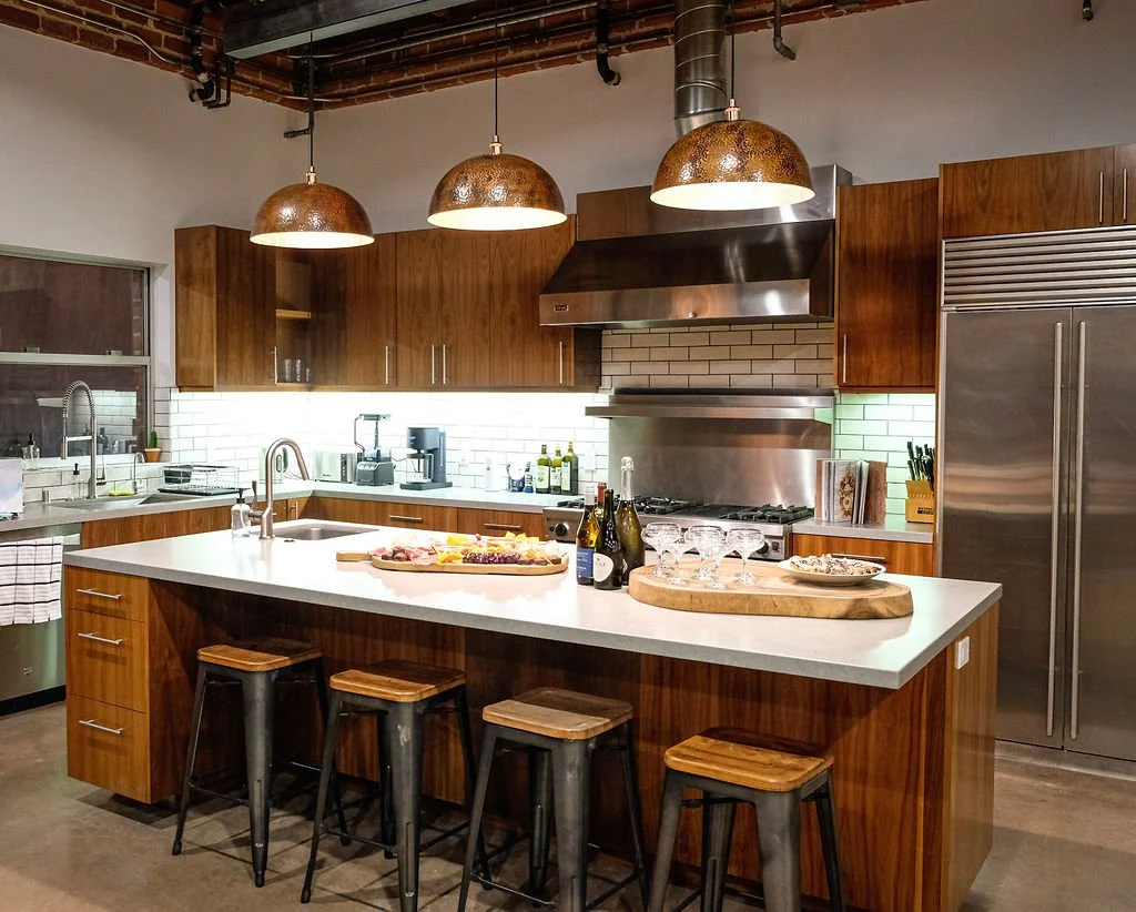 Modern kitchen with wooden cabinets, stainless steel appliances, white countertops, and three hanging pendant lights. There are four black and wood stools at the island, with a tray of drinks and snacks on top.