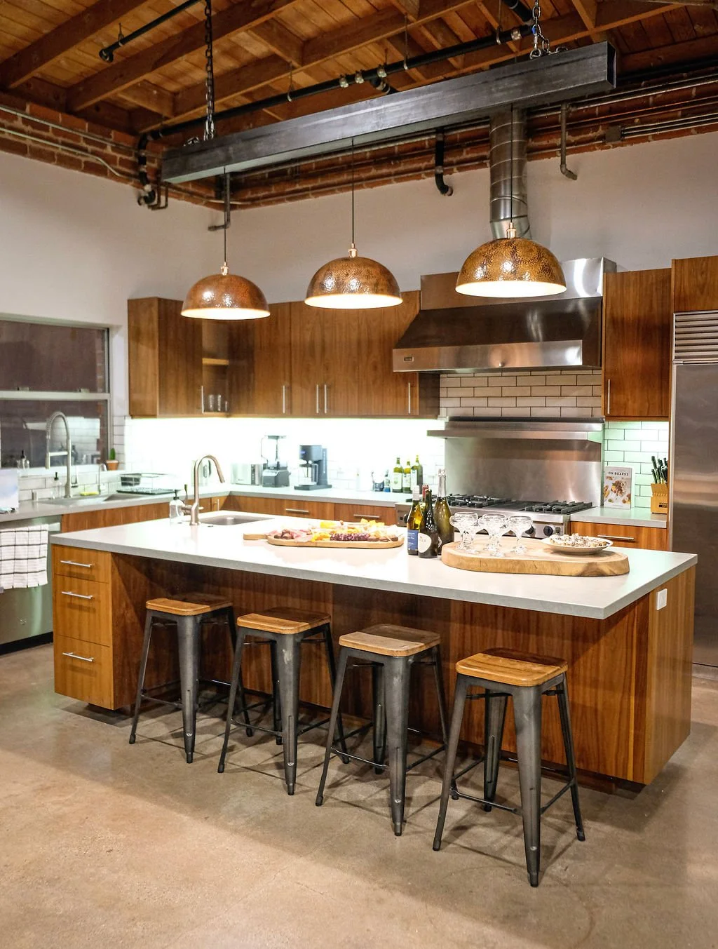 Modern kitchen with wooden cabinets, white countertops, and three hanging copper pendant lights. Contains a kitchen island with four stools, baking supplies, and kitchen appliances.