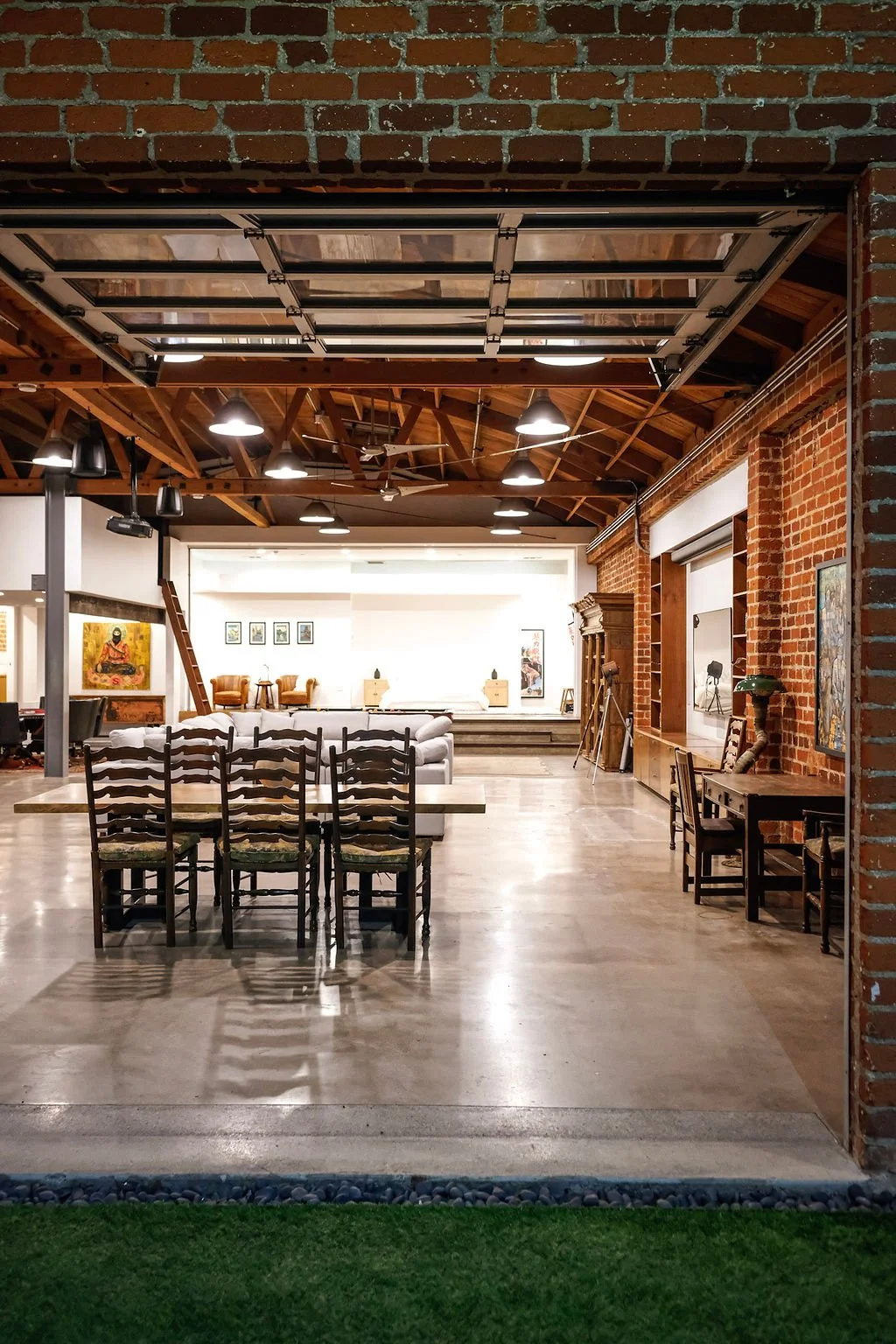 Interior view of an industrial-style living room with exposed brick walls, wooden ceiling beams, and modern lighting. The foreground features six wooden chairs arranged around a long dining table, with a white couch and artwork visible in the backgro