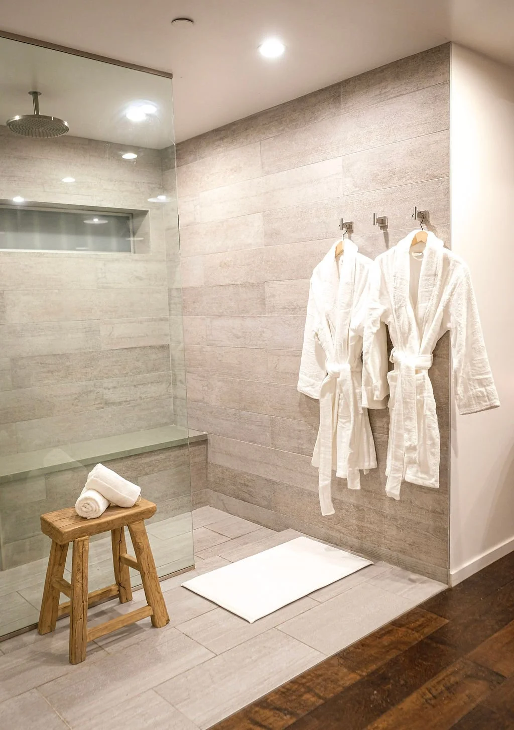Two white bathrobes hanging on hooks on a tiled wall in a bathroom or spa, with a wooden stool holding rolled white towels and a white rug on the floor.