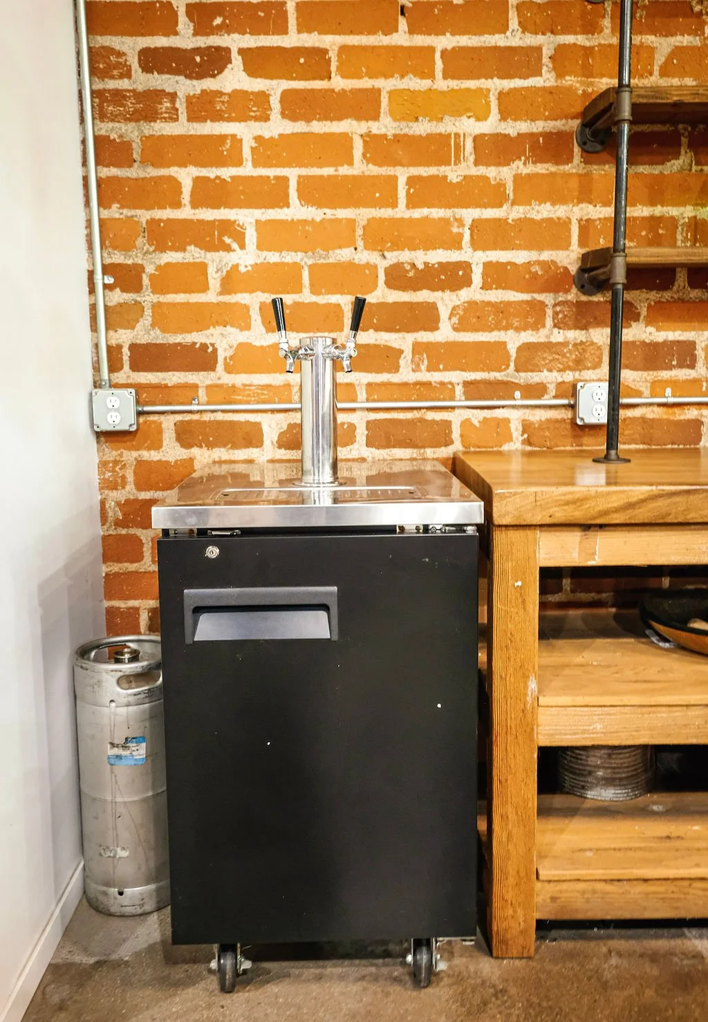 A beverage dispenser on a black rolling cabinet in a room with a brick wall, a wooden workbench to the right, and a gas cylinder to the left.