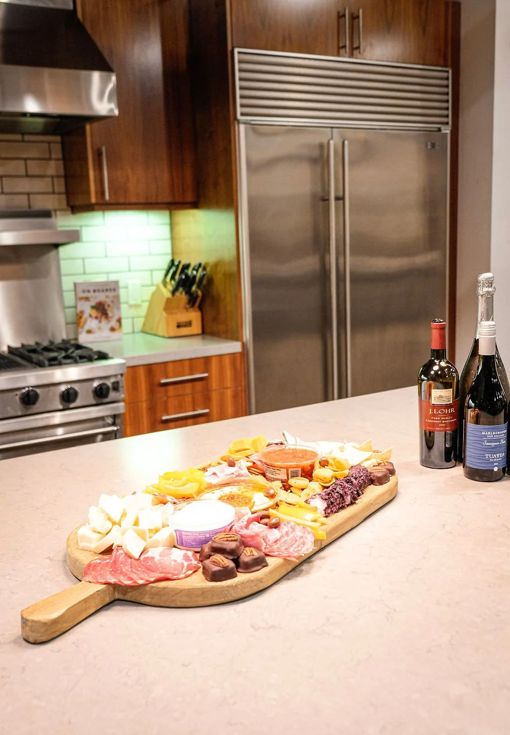 A wooden cutting board with assorted cheeses, meats, chocolates, and snacks on a kitchen counter, with two bottles of wine and a partner in the background.
