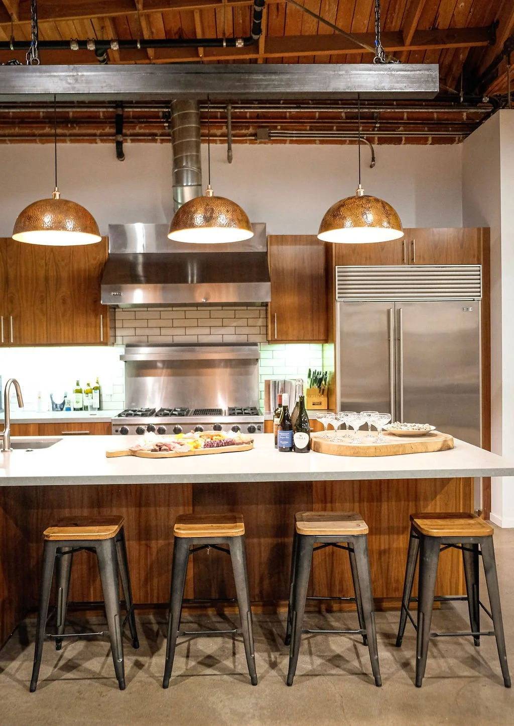 Modern kitchen with wooden cabinets, stainless steel appliances, pendant lights, and a white island counter with stools, wine bottles, and snacks.