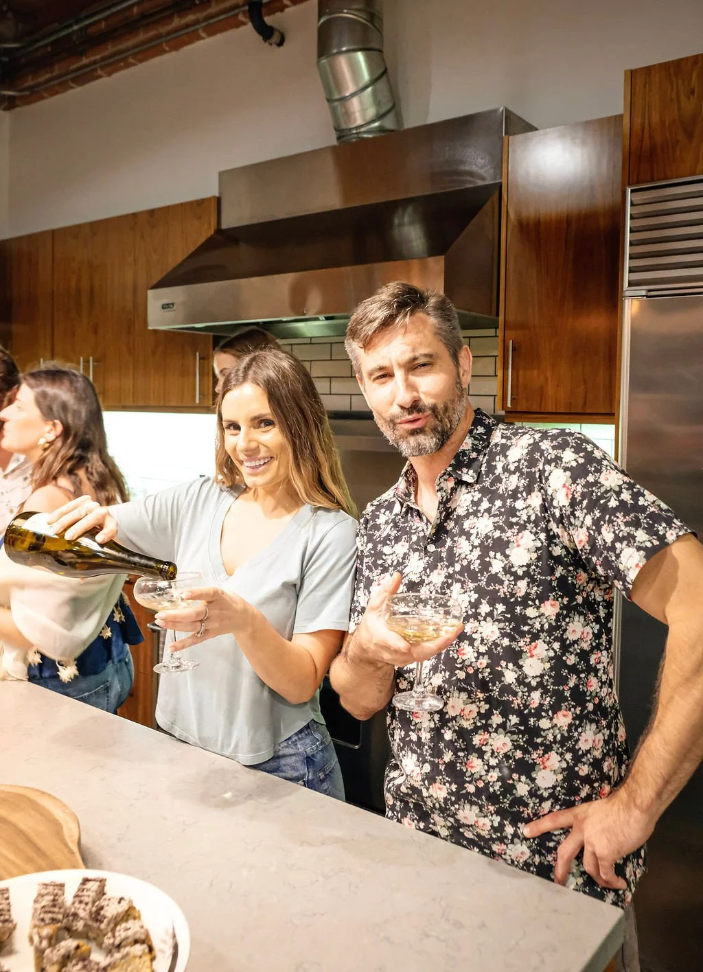 People enjoying a social gathering in a kitchen, with one woman pouring wine into a glass and a man holding a wine glass, while other people are in the background.