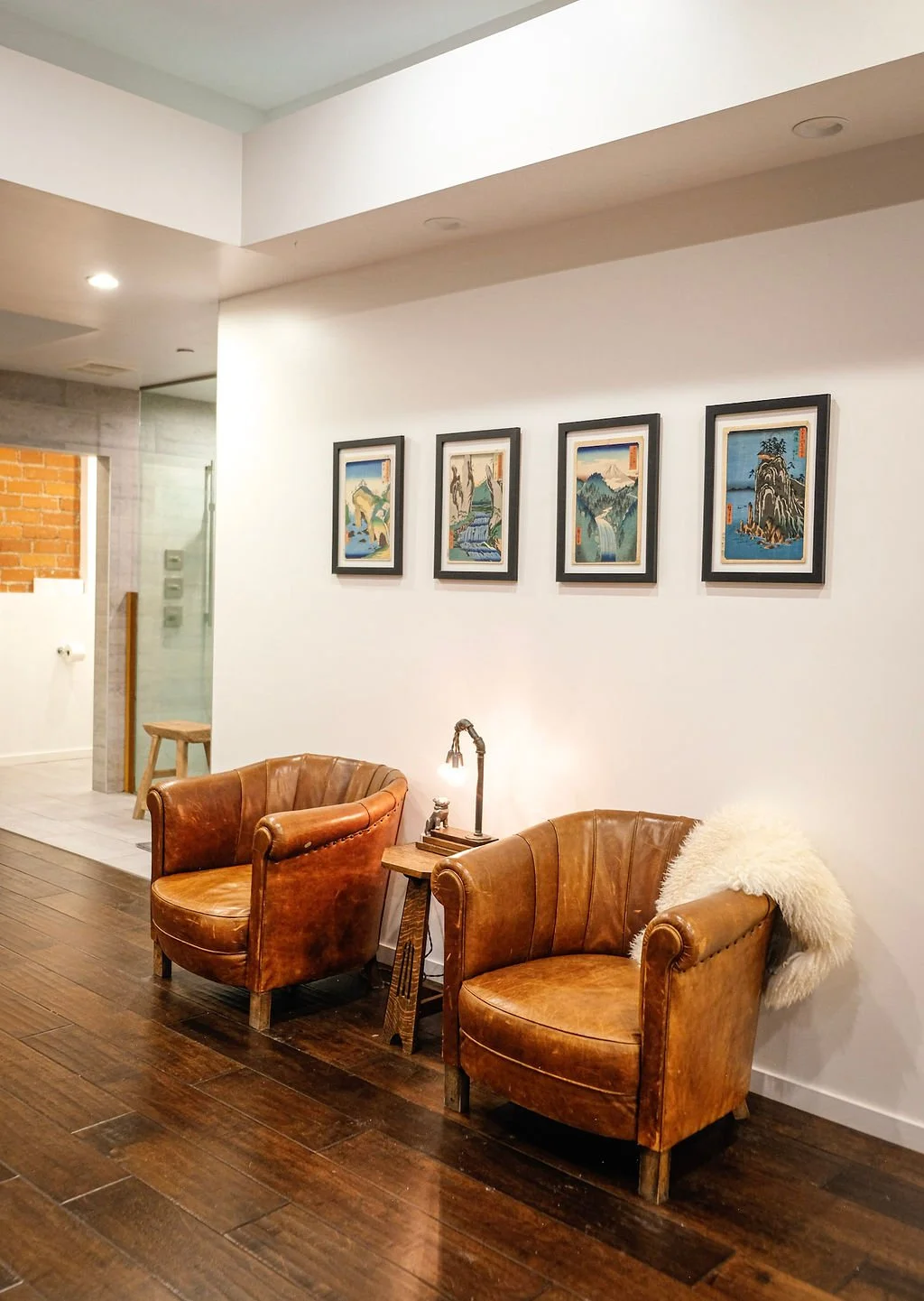 Two brown leather armchairs with a small wooden side table and a lamp in between, in a cozy interior with hardwood floors and decorative framed artwork of landscapes on a white wall.