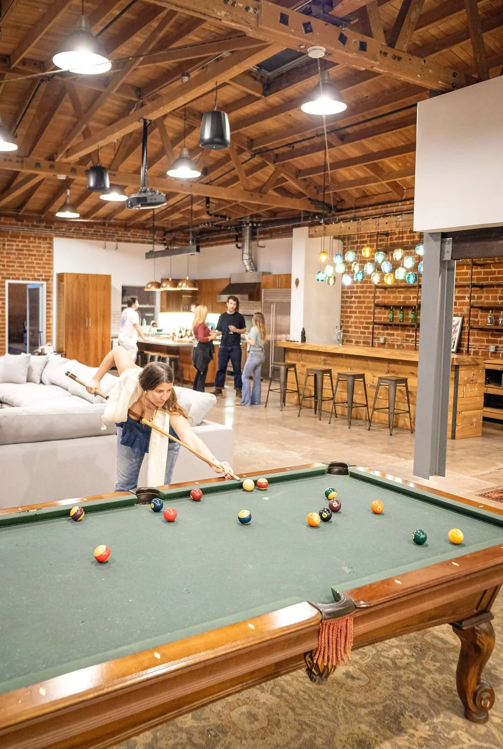 A woman is playing pool on a billiards table with a group of friends in a modern, rustic-style basement with exposed brick walls, wooden ceiling beams, and warm lighting.