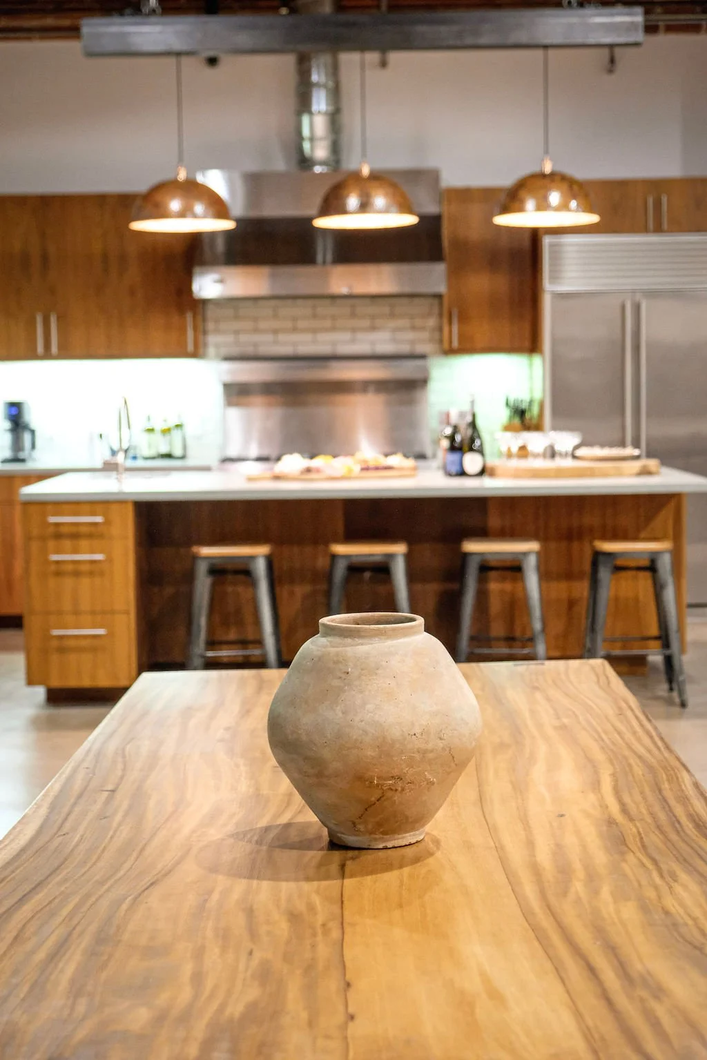 A rustic kitchen with a clay vase on a wooden table, wooden cabinets, a kitchen island with stools, stainless steel appliances, and copper pendant lights.