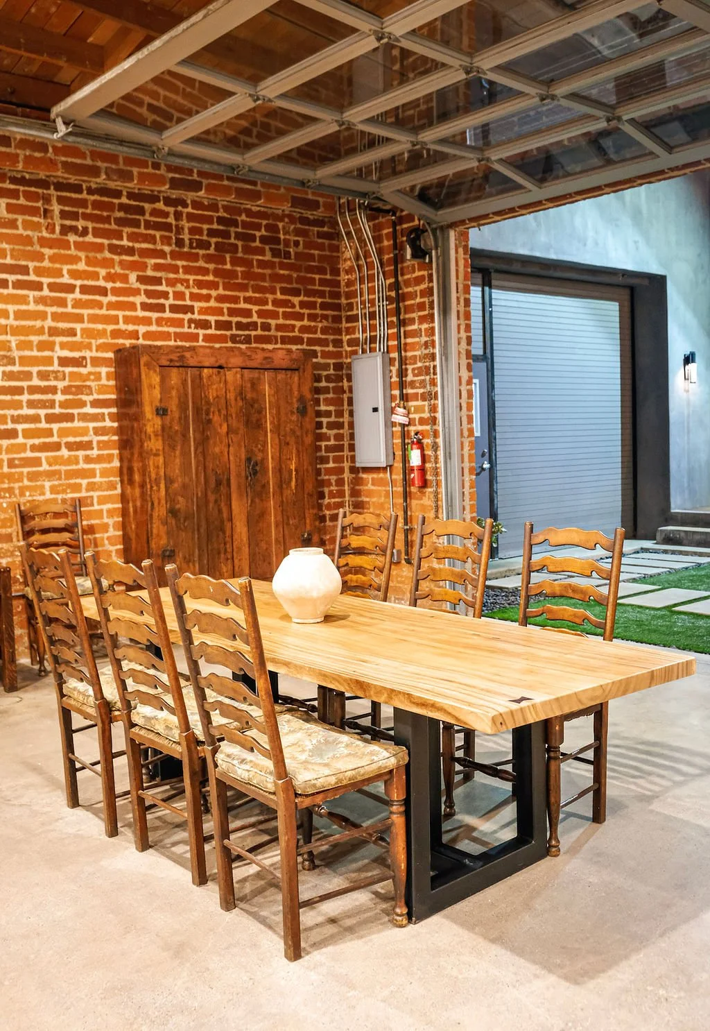 Indoor dining area with a rustic wooden table surrounded by eight wooden chairs with woven seats, against a brick wall. A white vase is on the table. Part of a garage door and door with stairs outside are visible. The ceiling is wooden with metal fra