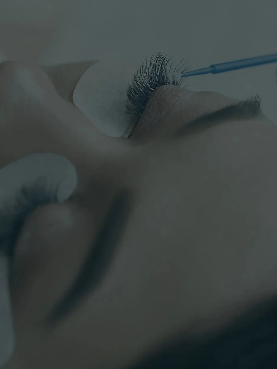 Close-up of someone receiving eyelash extensions, with a technician applying adhesive to natural eyelashes using a small brush.