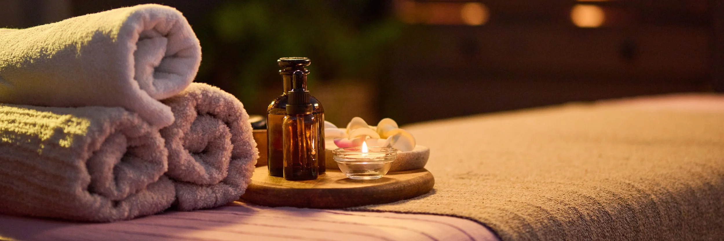 Spa setting with rolled towels, essential oil bottles, candle, and flower petals on a wooden tray, softly lit for relaxation.