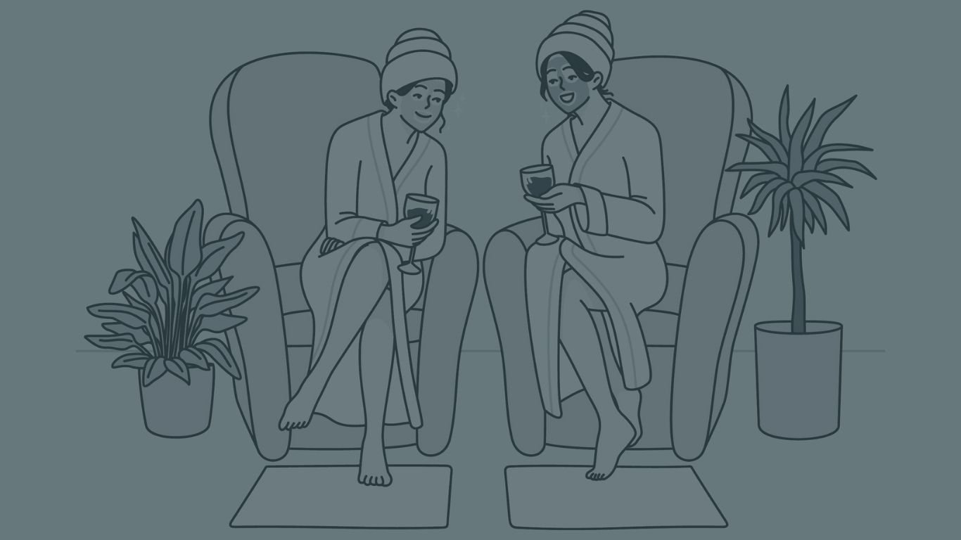 Two women sitting on armchairs, wearing robes and towels on their heads, holding drinks and smiling, with potted plants beside them in a cozy room.
