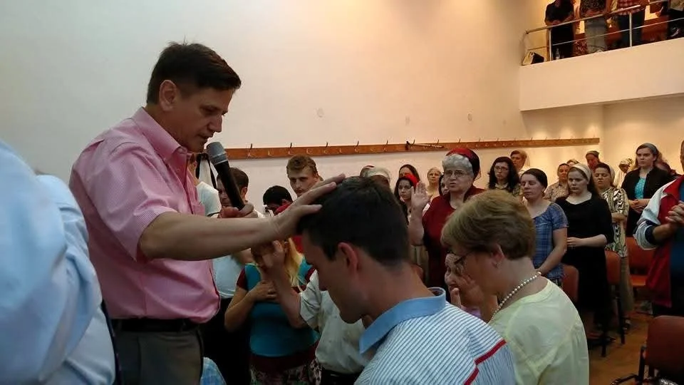 A man in a pink shirt is performing a religious or spiritual ritual by placing his hand on the head of a young man with dark hair, during an indoor gathering with many people, some standing and some sitting, some wearing Santa hats.