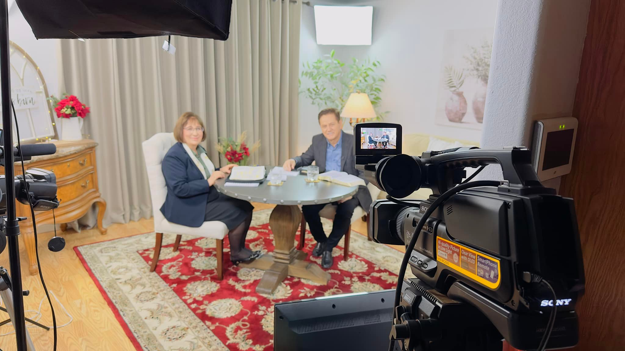 Two people sitting at a round table during a video recording or interview in a cozy, well-lit room with plants, artwork, and a colorful rug.