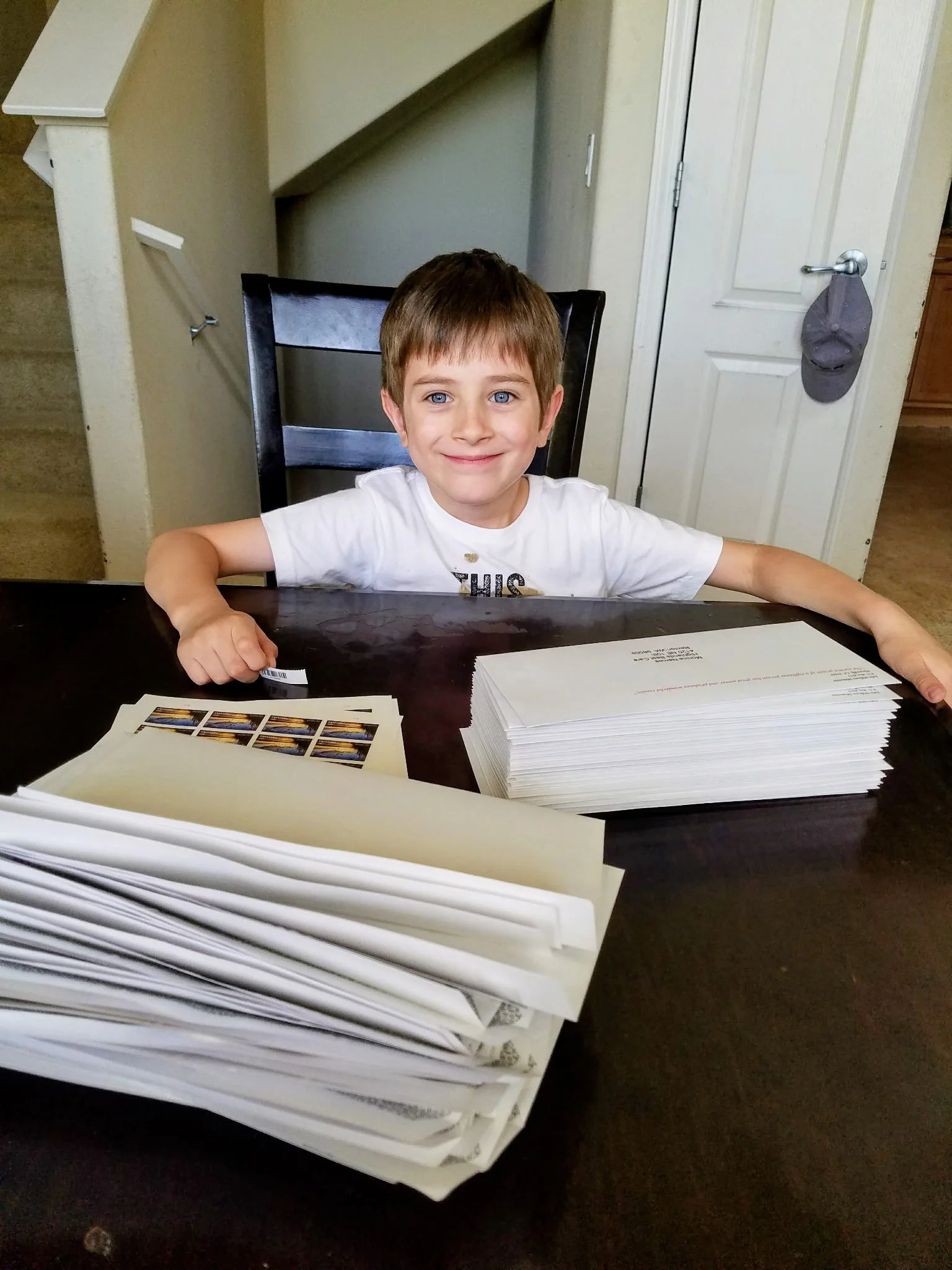 A young boy with brown hair and blue eyes sitting at a dark wooden table, smiling at the camera. In front of him are multiple stacks of printed papers or envelopes, some with stamps, arranged on the table. The background shows a staircase, a white door with a gray cap hanging on the door handle, and part of a kitchen area.