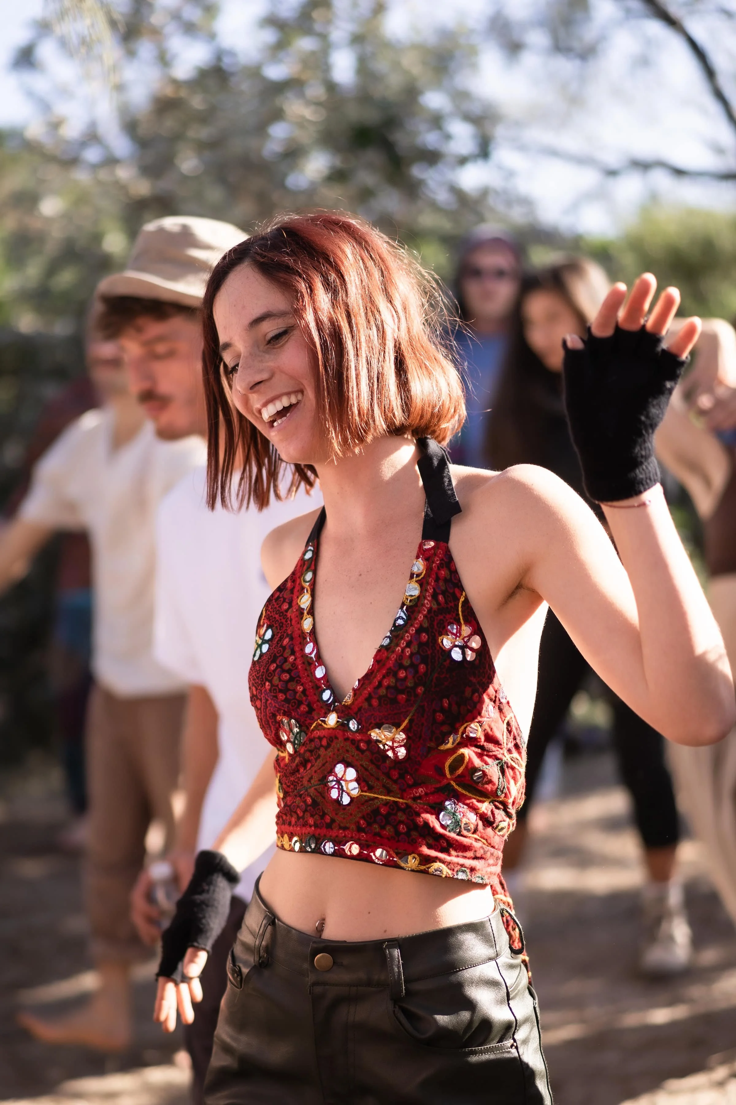A young woman with short red hair, wearing a red floral crop top, black pants, and fingerless gloves, smiling and waving with her right hand, in an outdoor setting with other people in the background.