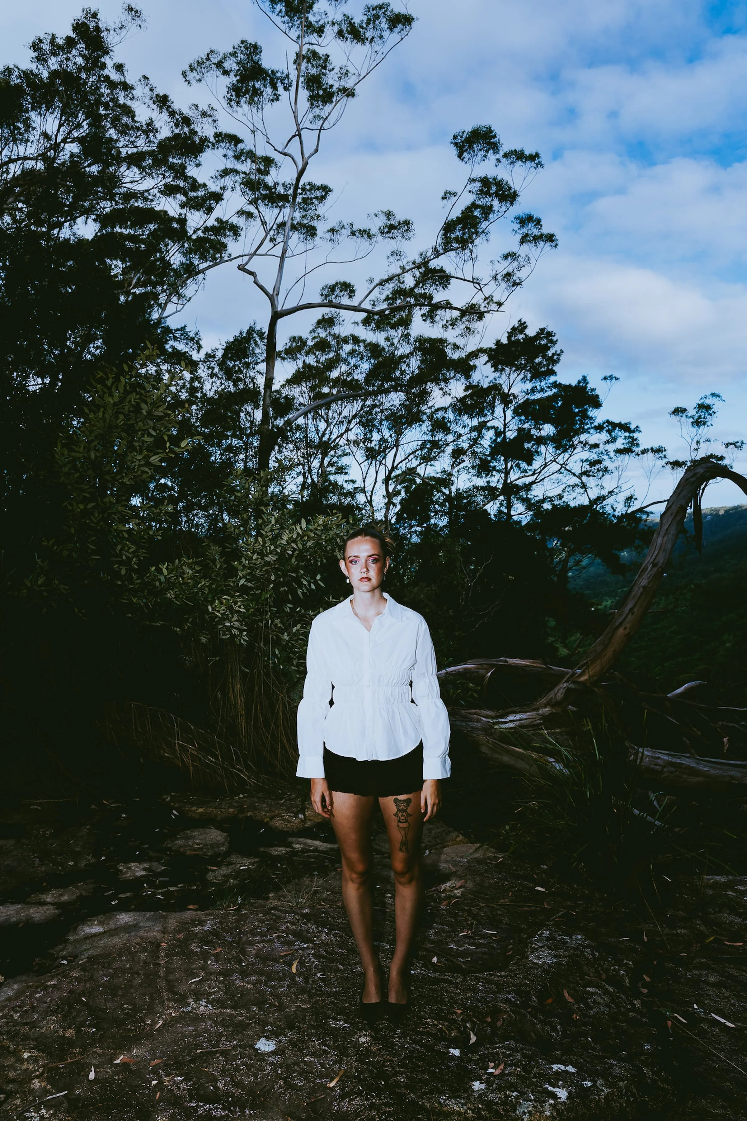 A woman standing outdoors in a wooded area with a cloudy sky background, wearing a white top, black shorts, and black high heels.