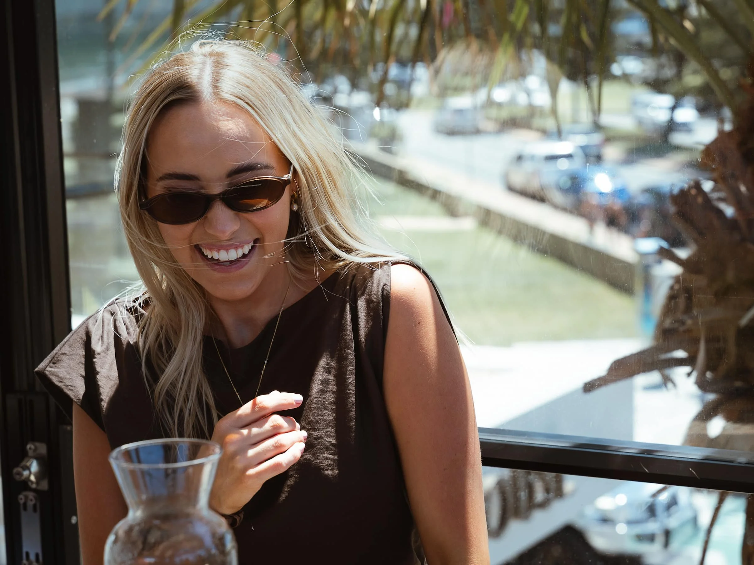 A blonde woman wearing sunglasses and a dark sleeveless top is smiling and laughing inside a vehicle or building near a window, with cars and trees visible outside.