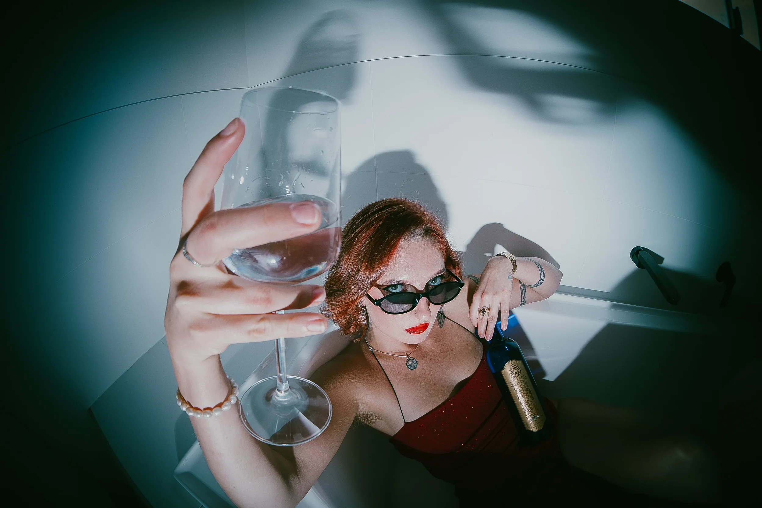 Person with red hair and sunglasses lying in a bathtub, holding a wine glass and a bottle of wine, with a moody, artistic lighting.