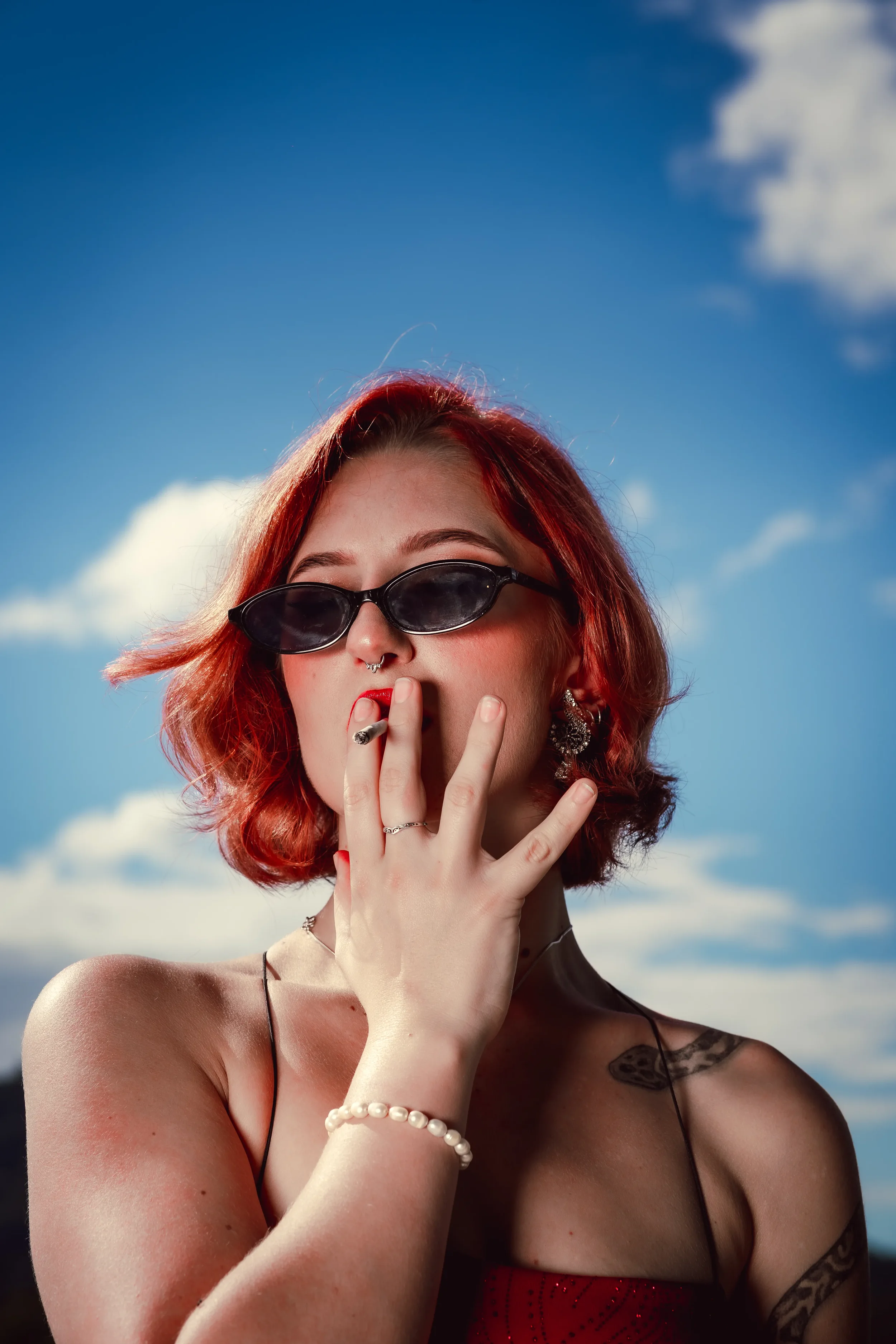 A woman with red hair and sunglasses smoking a cigarette against a bright blue sky with a few clouds.