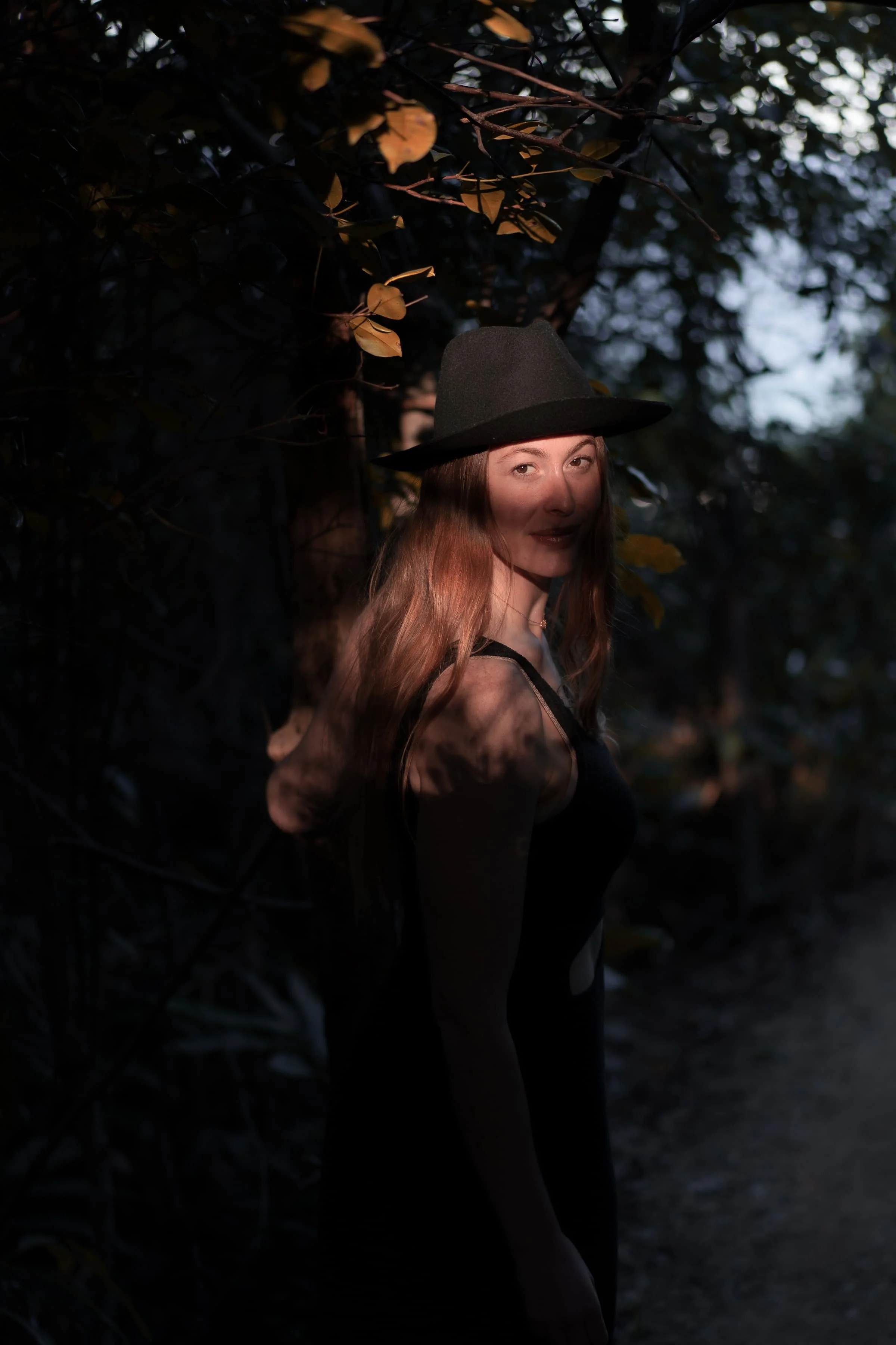 A young woman with long red hair wearing a dark sleeveless top and a wide-brimmed hat stands against a tree in a forest, with dappled sunlight highlighting her face.