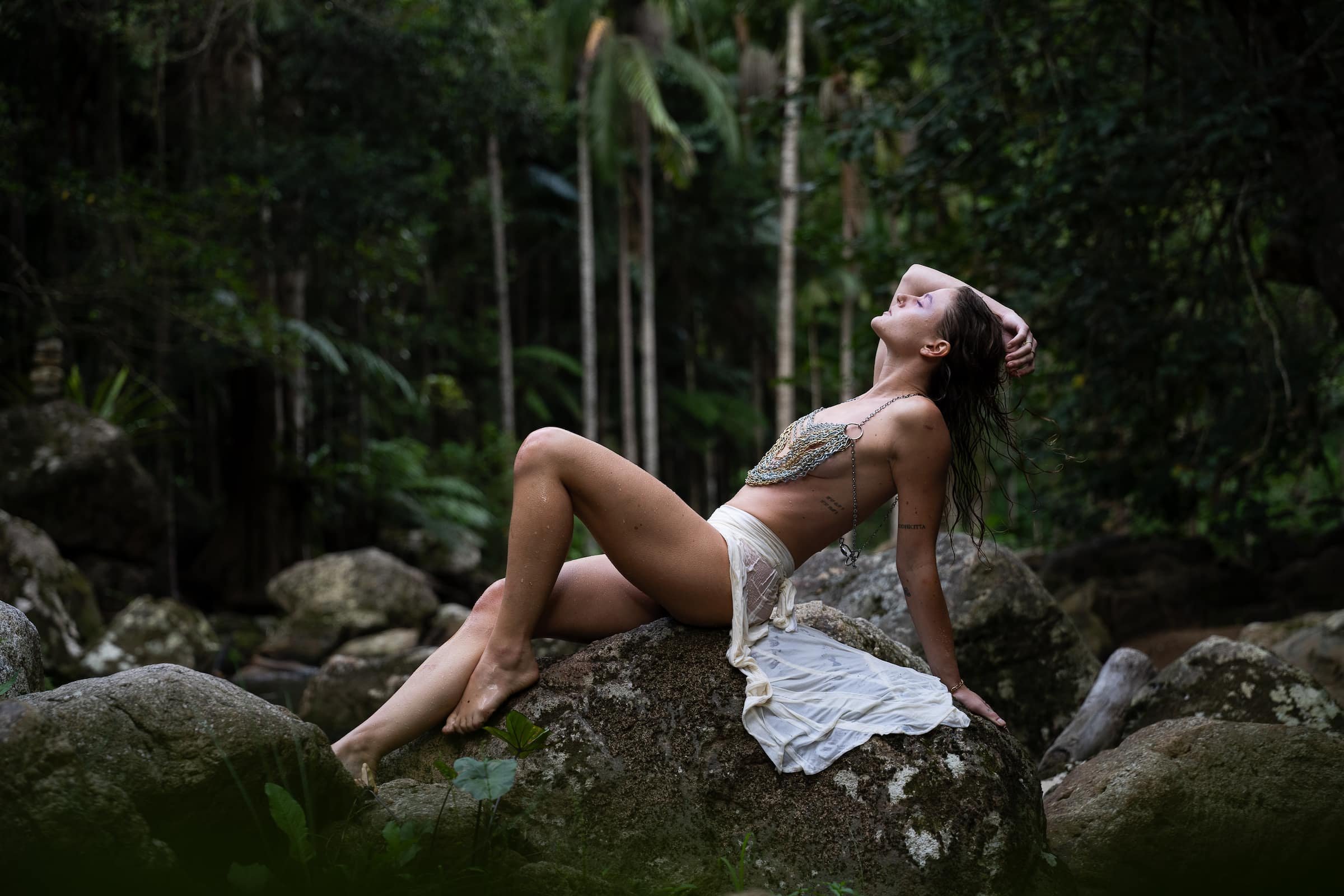 A woman with long dark hair and tattoos reclining on a large rock in a lush, green forest, with her head tilted back and eyes closed.