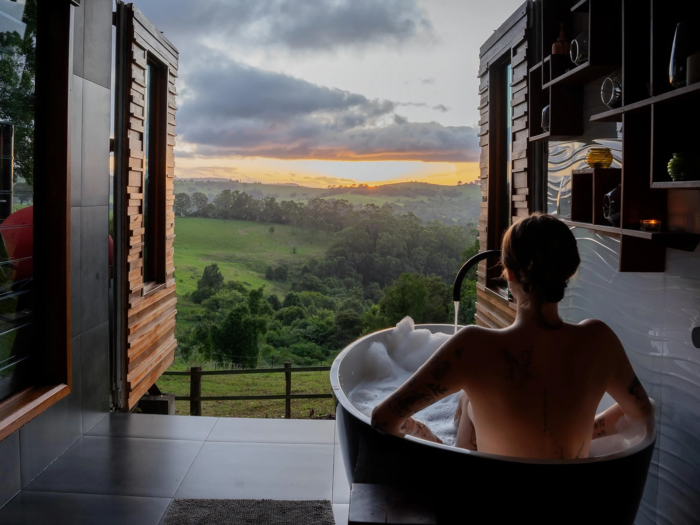 A person with tattoos soaking in a bubble-filled bathtub, looking out through open windows at a scenic view of green rolling hills and a sunset sky with clouds.