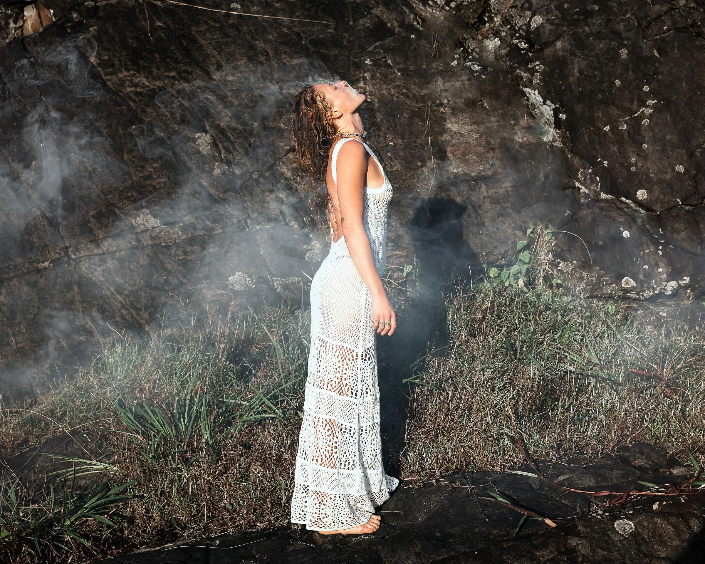 A woman in a white crocheted dress standing barefoot outdoors near rocks and grass, with her head tilted back and smoke or mist surrounding her.