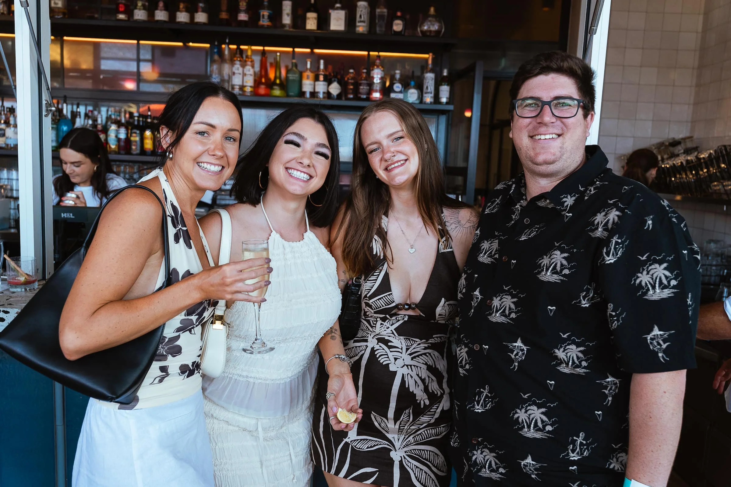 Four friends smiling at a bar, holding drinks, with shelves of liquor bottles in the background.