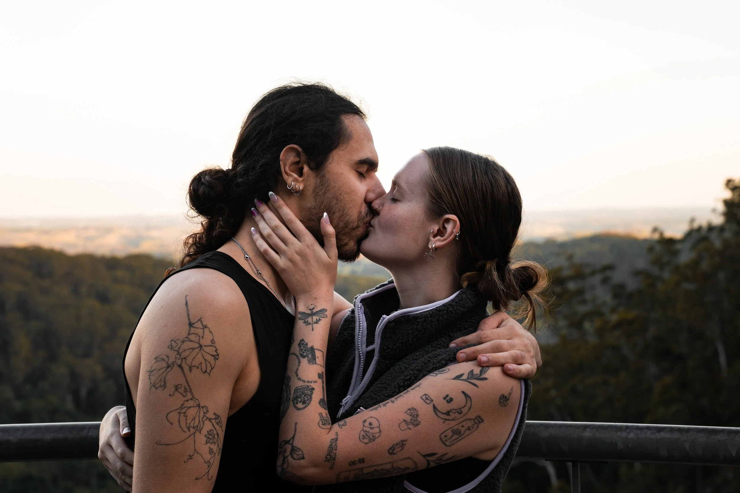 A couple sharing a kiss outdoors, with a scenic landscape in the background. They have tattoos on their arms and are holding each other closely.