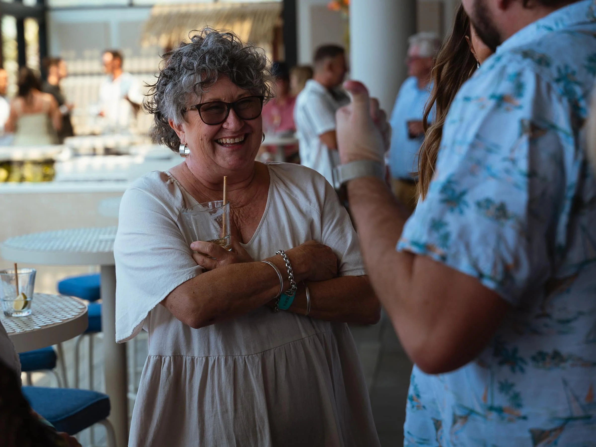 A cheerful older woman with glasses and curly gray hair smiling while holding a drink, engaged in a conversation with a man in a floral shirt at a lively indoor social gathering.