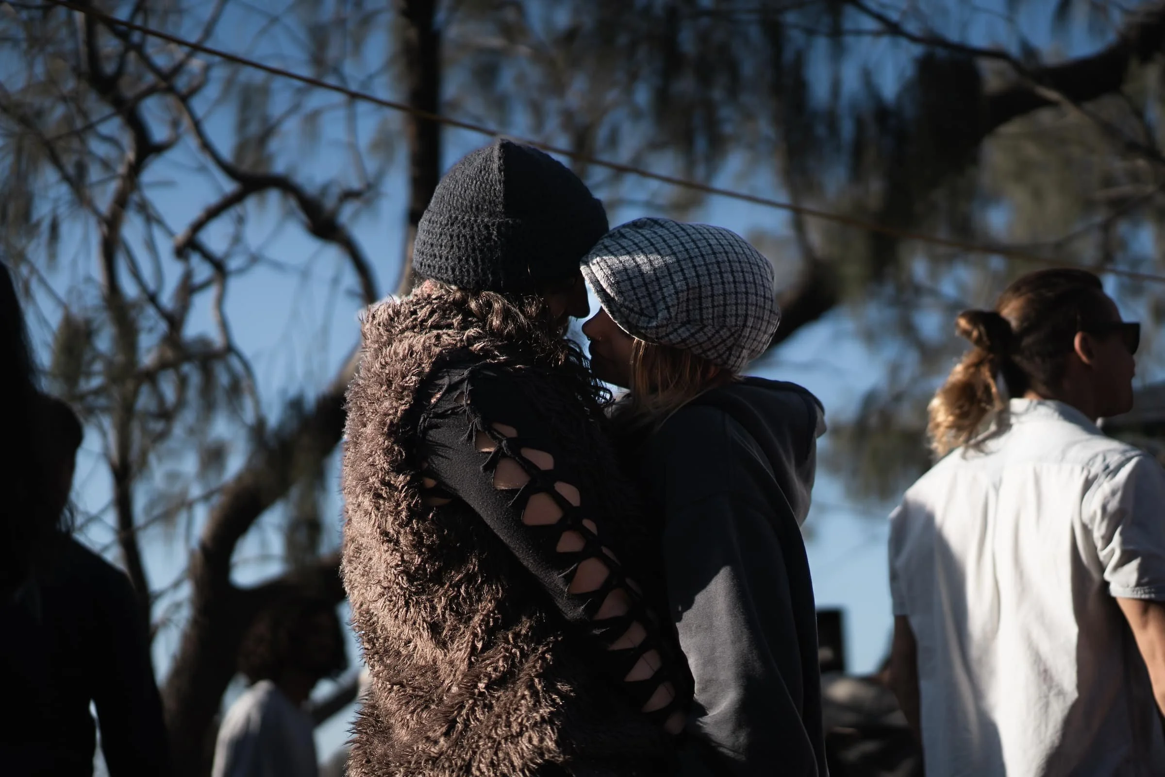 Two women with winter hats and jackets touching foreheads in an outdoor setting with trees, while other people are in the background.
