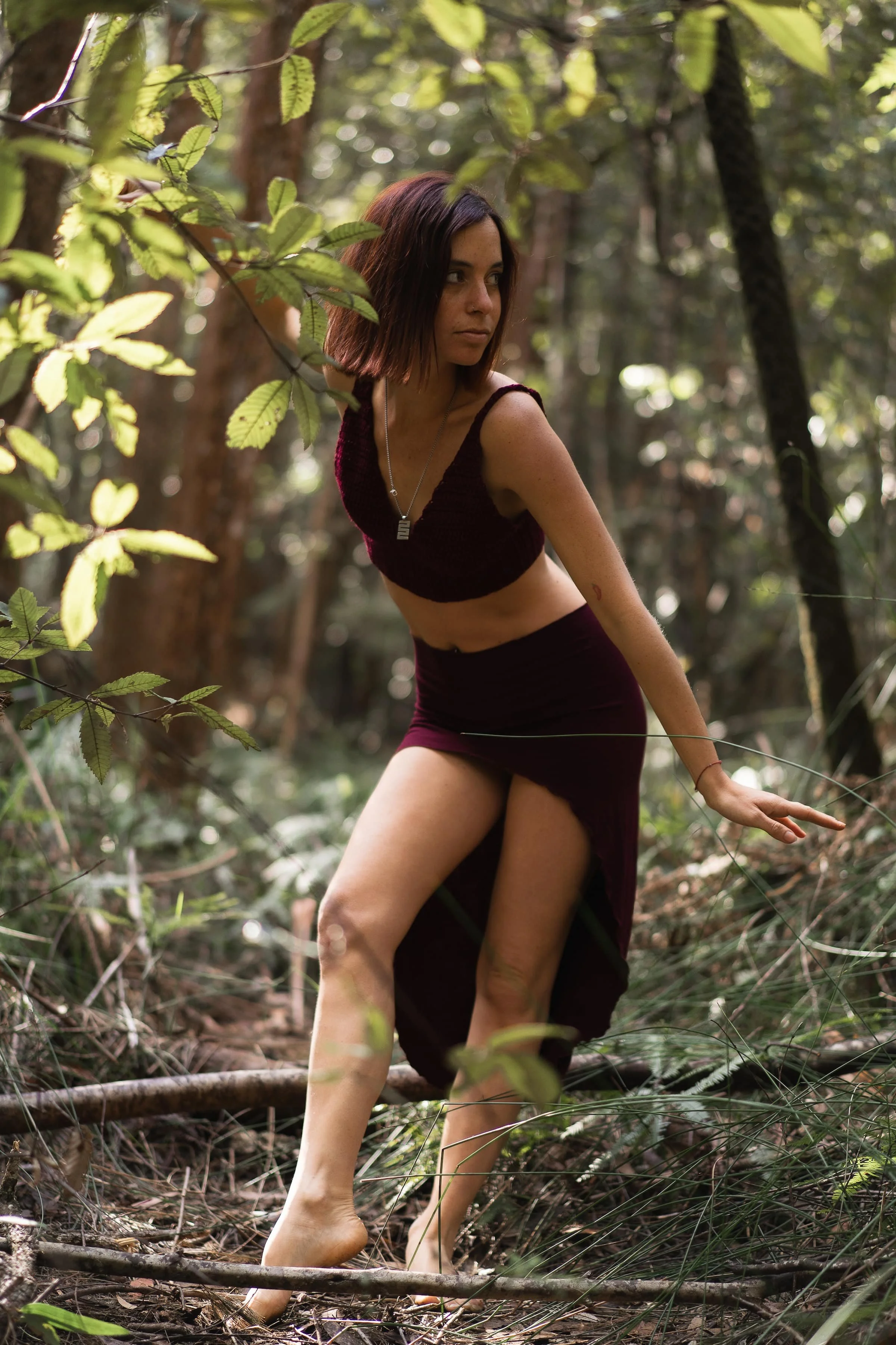 A young woman with shoulder-length dark hair wearing a dark purple crop top and skirts, walking barefoot through a lush forest with dense greenery and trees.