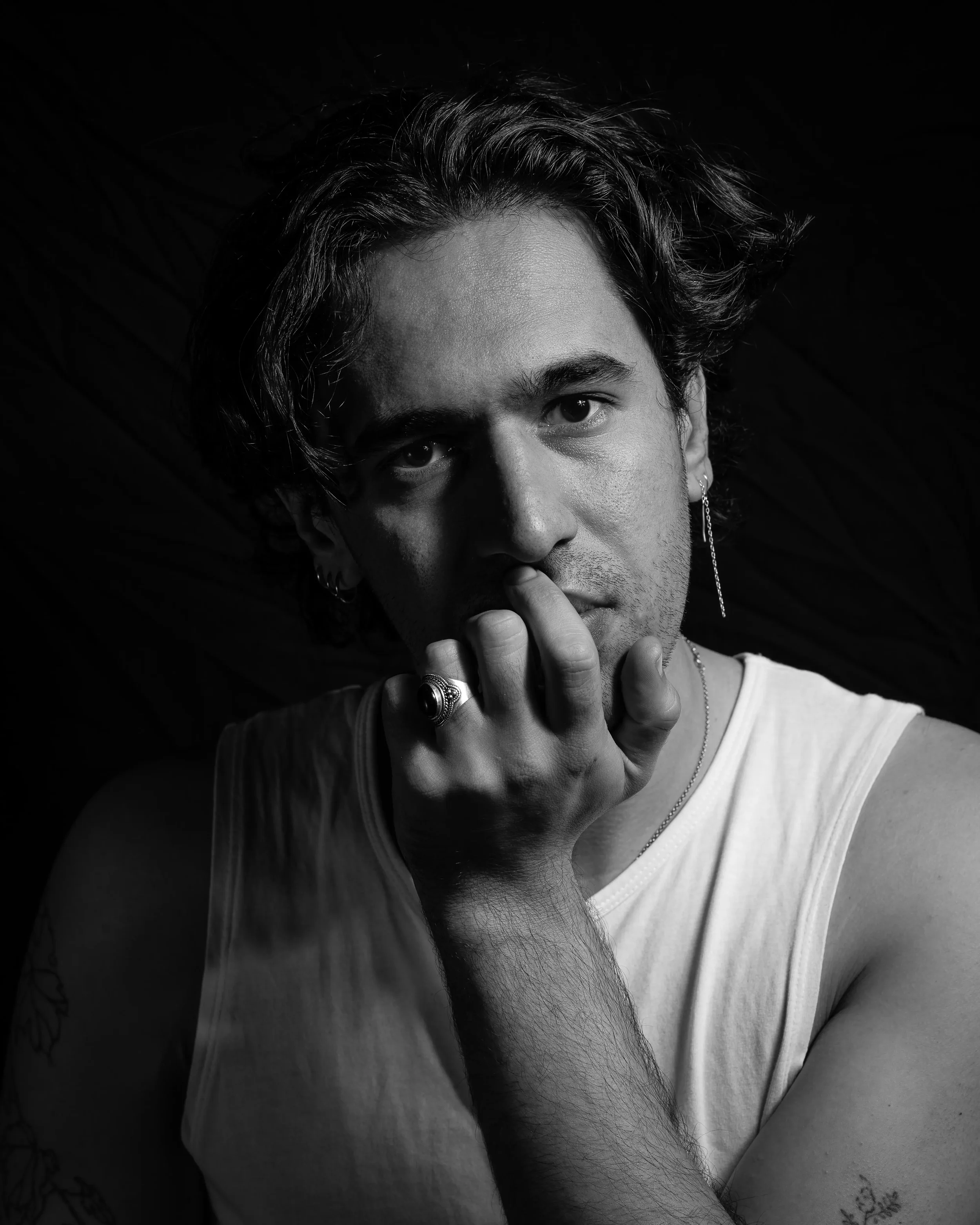 Black and white portrait of a young man with dark hair, multiple earrings, wearing rings and a necklace, with a contemplative expression and hand near his chin.