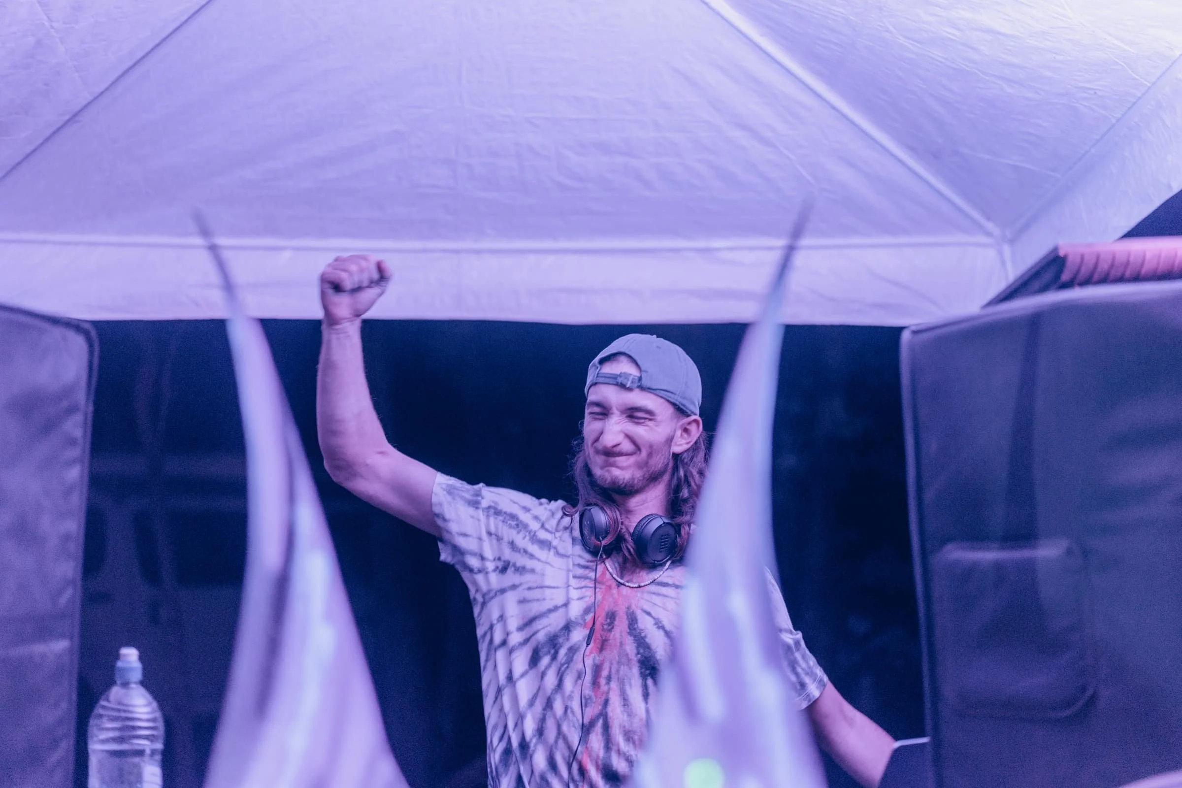 A man with long hair, wearing a cap backwards and headphones around his neck, dances with a fist in the air under a tent with purple lighting.