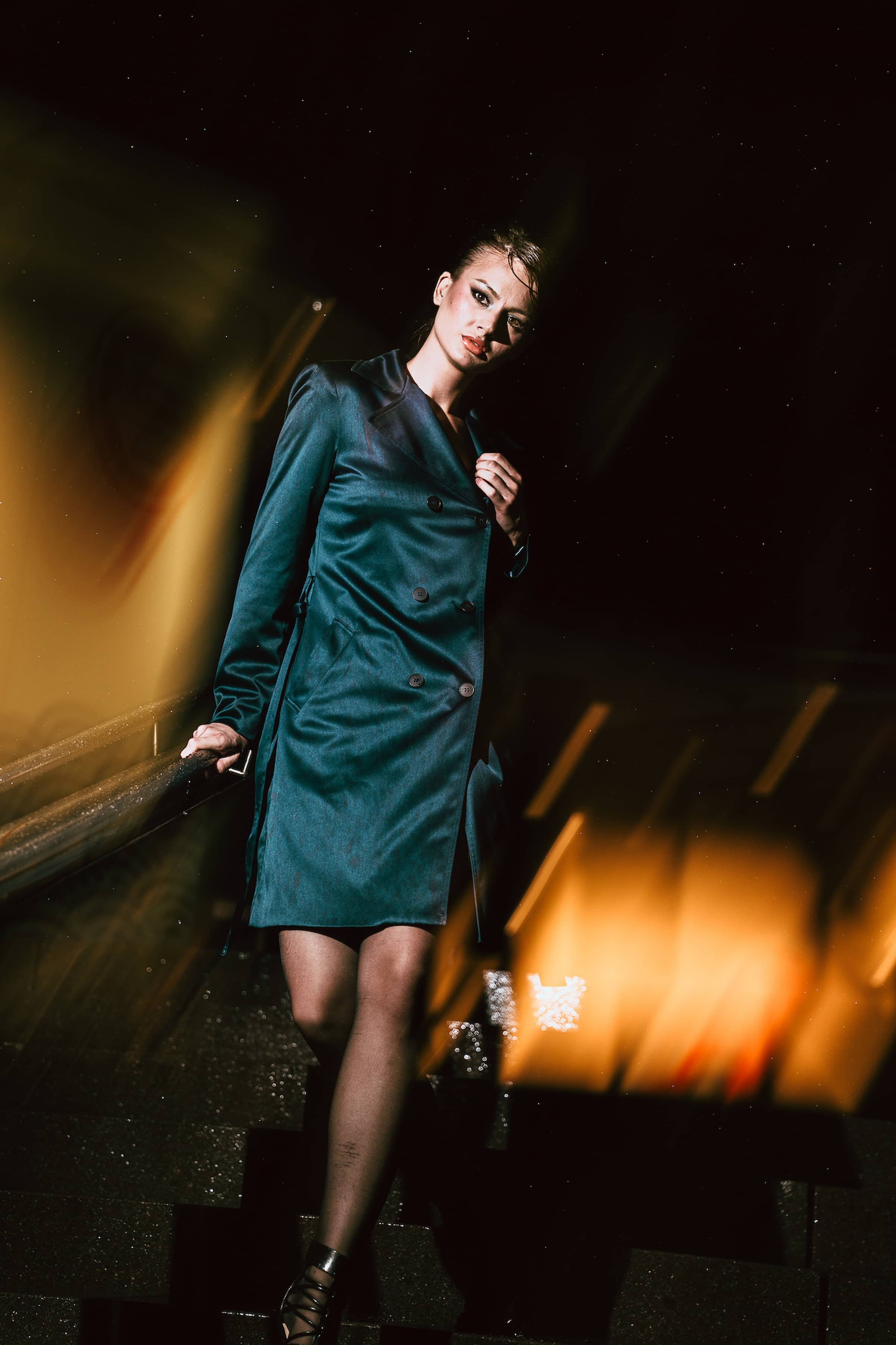 A woman in a teal satin trench coat and black lace-up high heels stands on a wet street at night under a starry sky with orange and yellow lighting effects in the background.