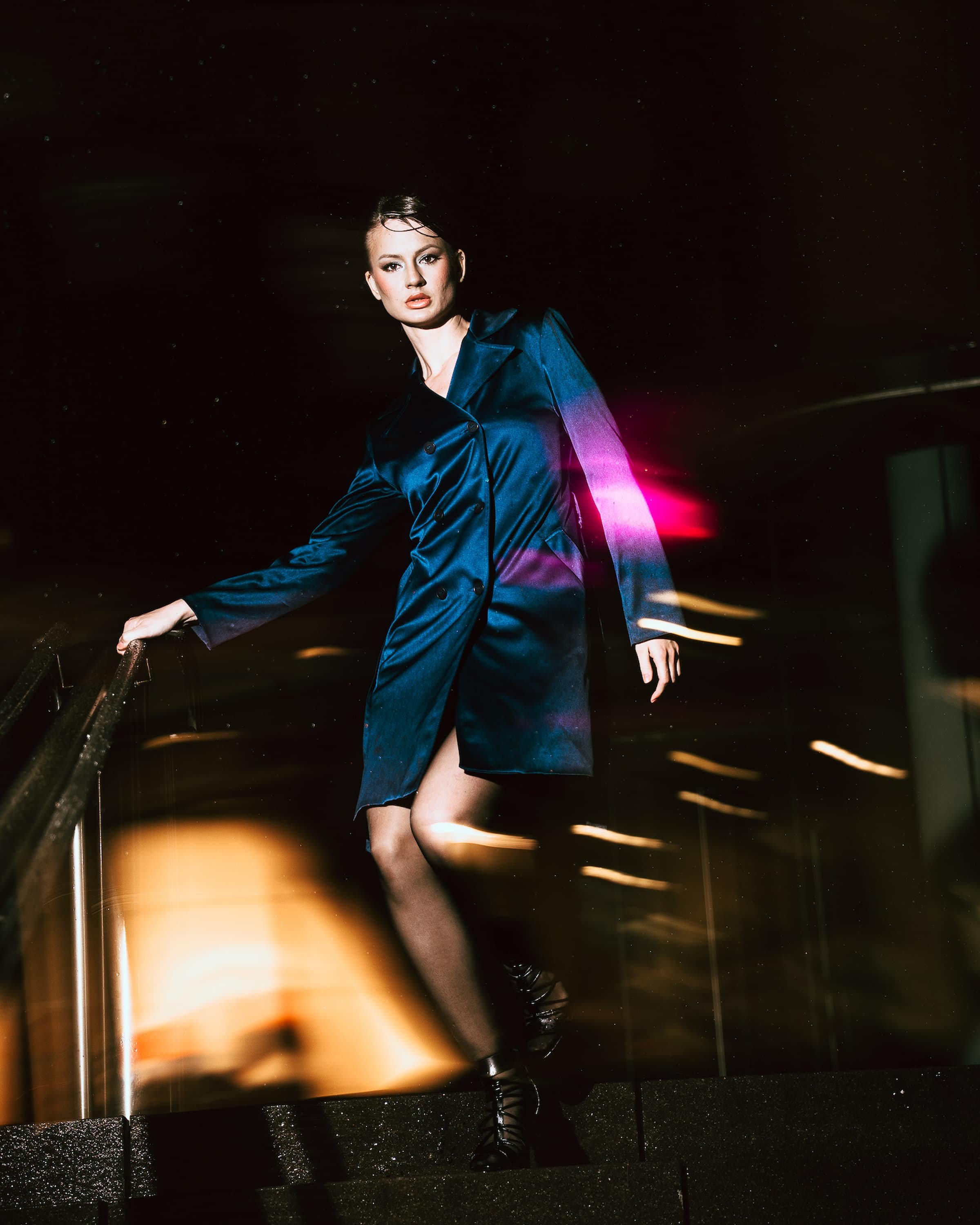 A woman in a dark blue trench coat walking down a staircase at night with city lights and colorful light streaks in the background.