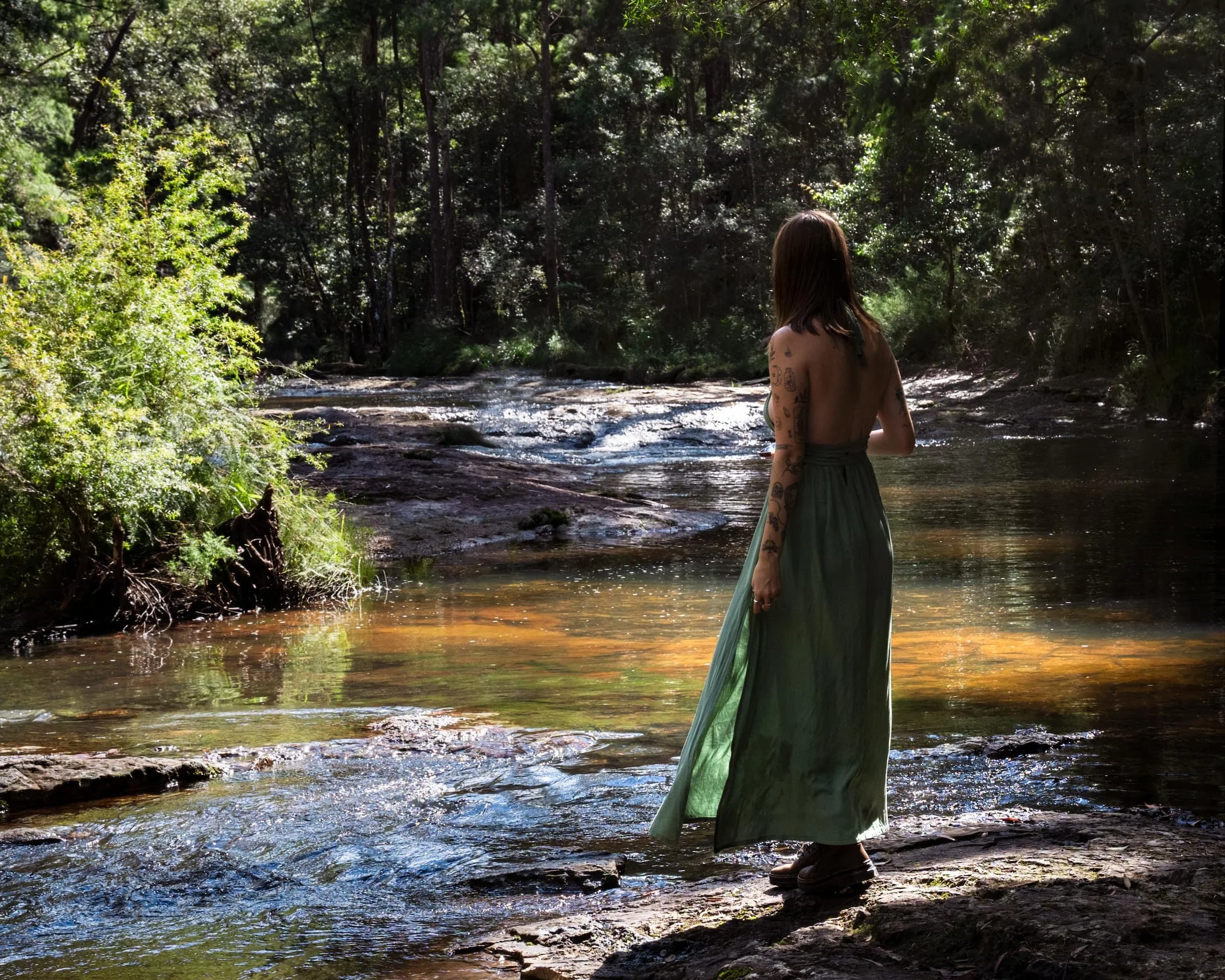 Woman with tattoos in a green skirt standing on rocks at the edge of a river in a lush forest during daytime.