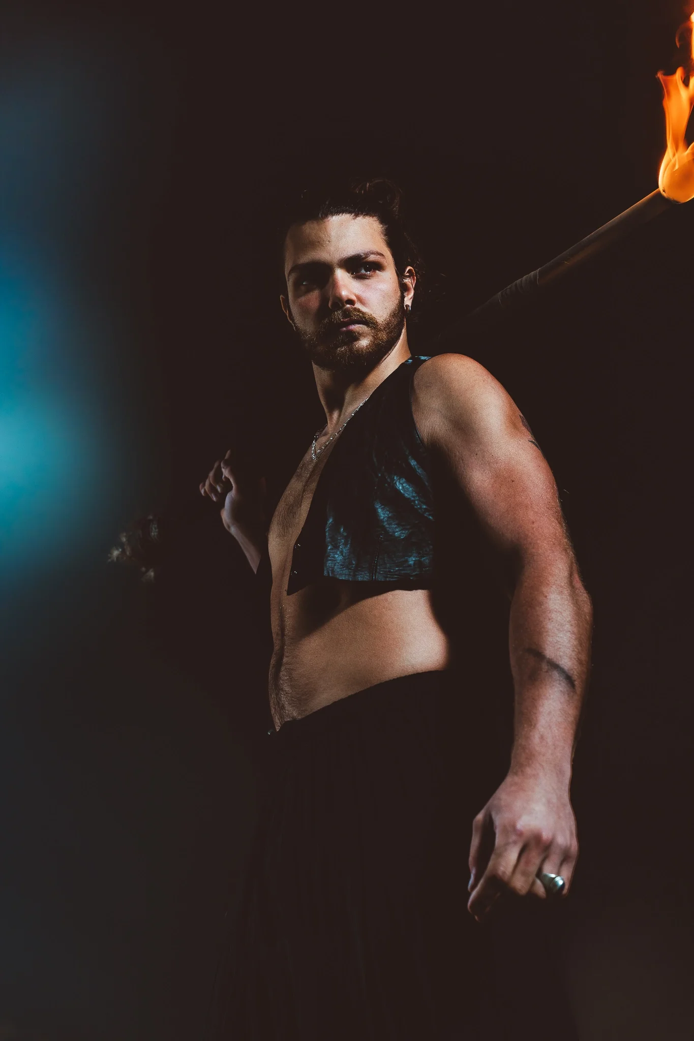 A person with dark hair and a beard looking over their shoulder. They are wearing a dark, sleeveless vest and black pants. The background is black with a bright blue light on the left and a torch with flame on the right.