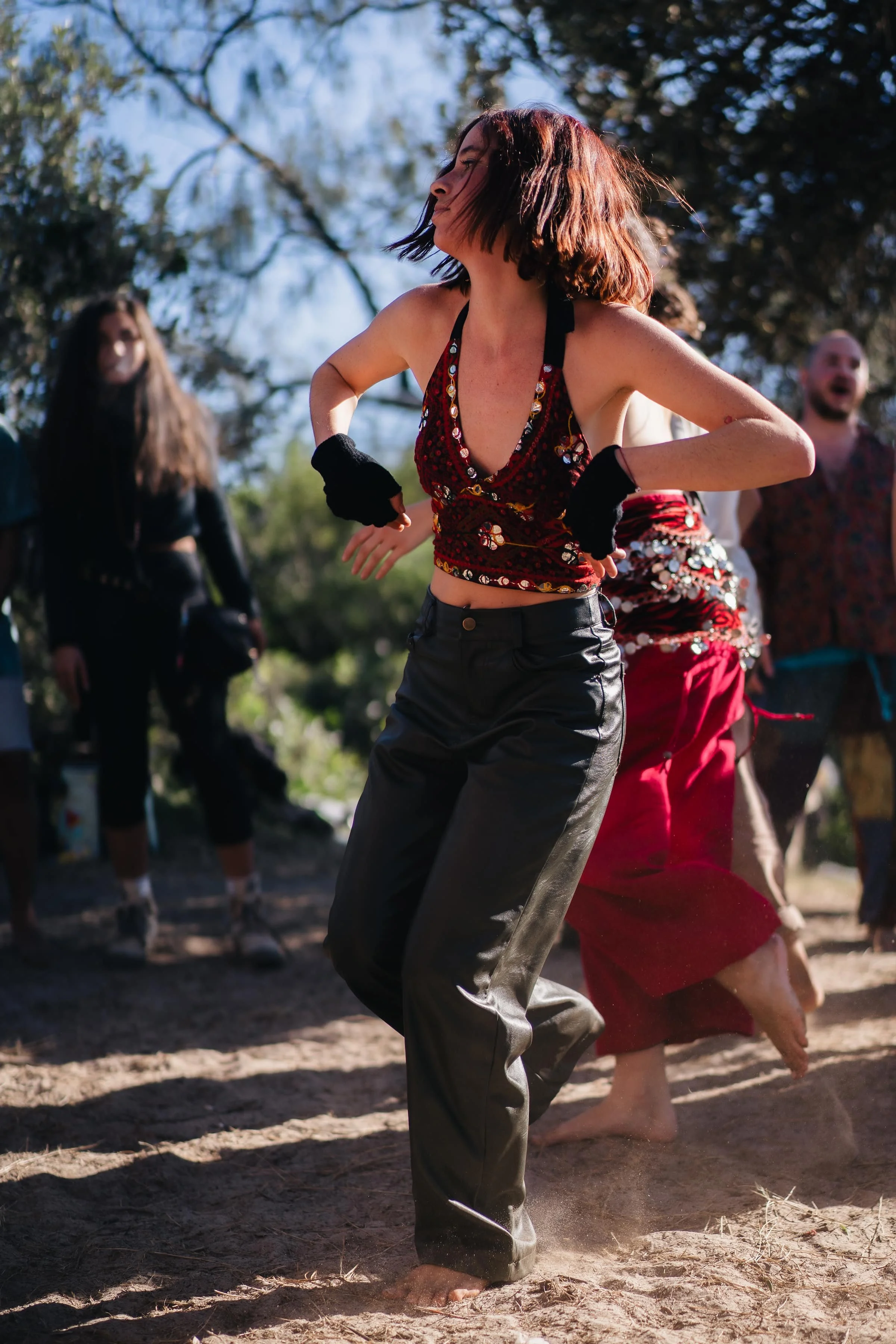 A woman dancing outdoors on dirt ground, surrounded by other people, with trees and blue sky in the background.