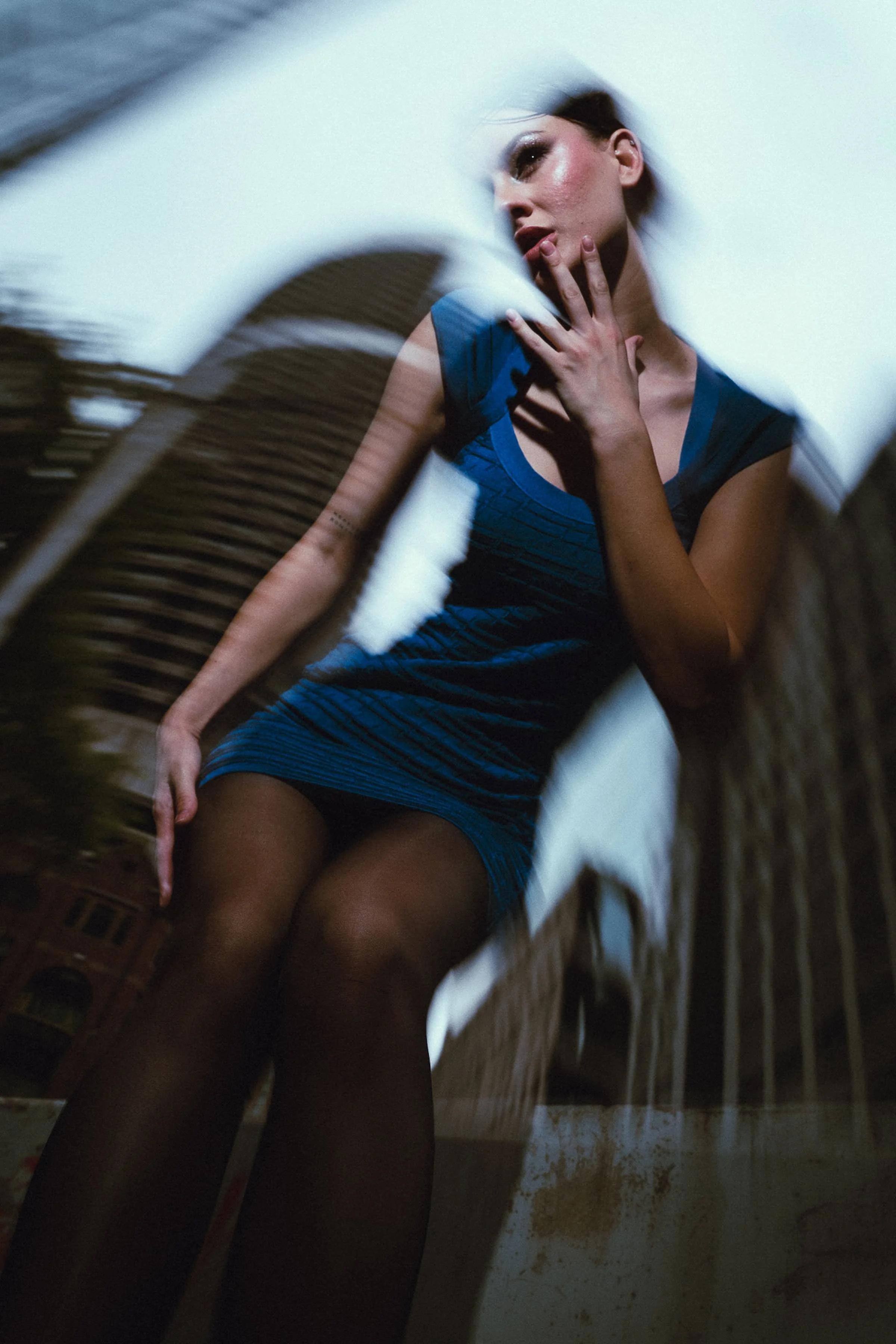 A woman in a blue dress posing with her hand near her face, shot through a blurred, circular object.