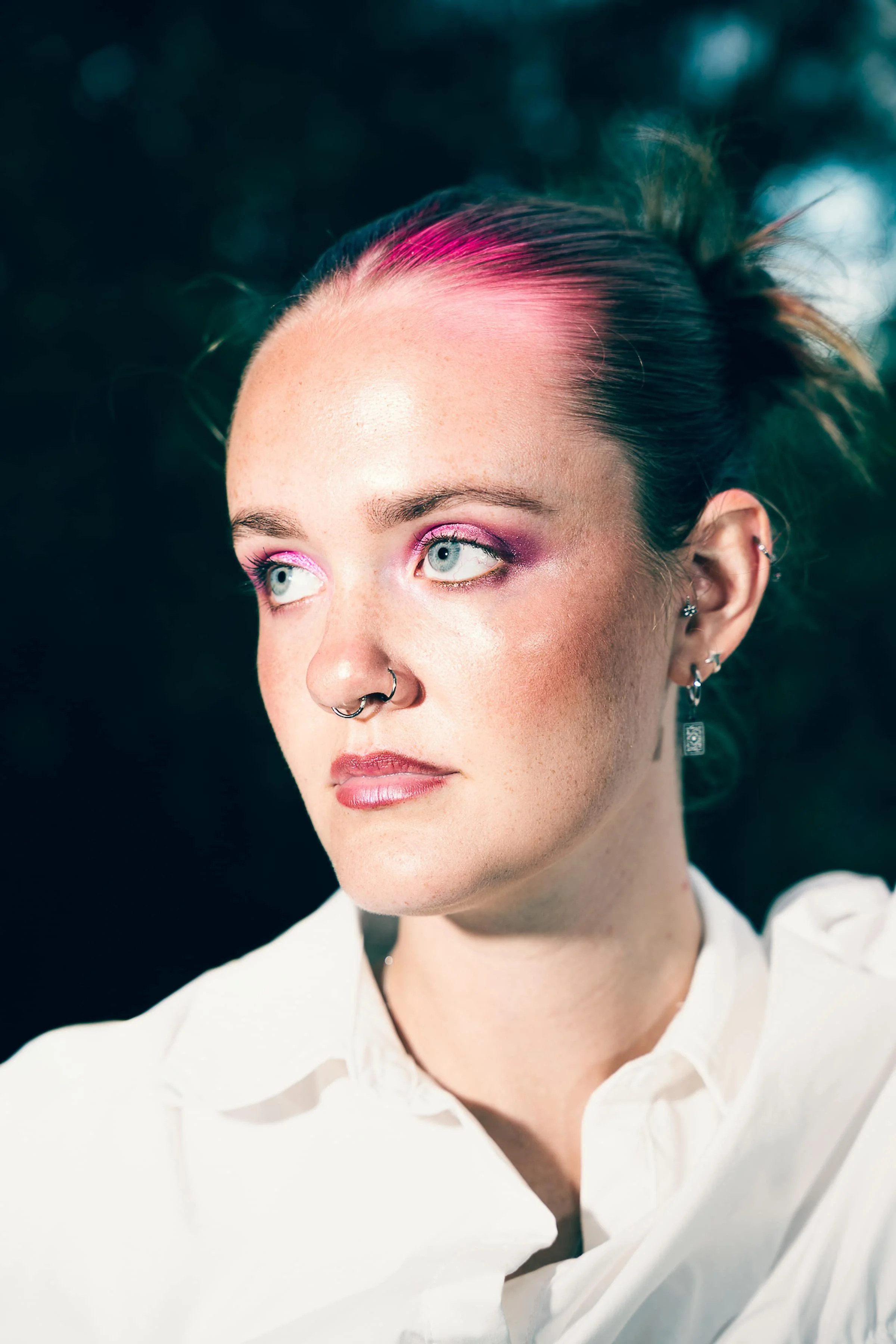 A woman with pale skin, pink hair streaks, and blue eyes looking to the side, wearing earrings, with makeup that includes pink eyeshadow and lipstick, in a white collared shirt against a dark blurred background.