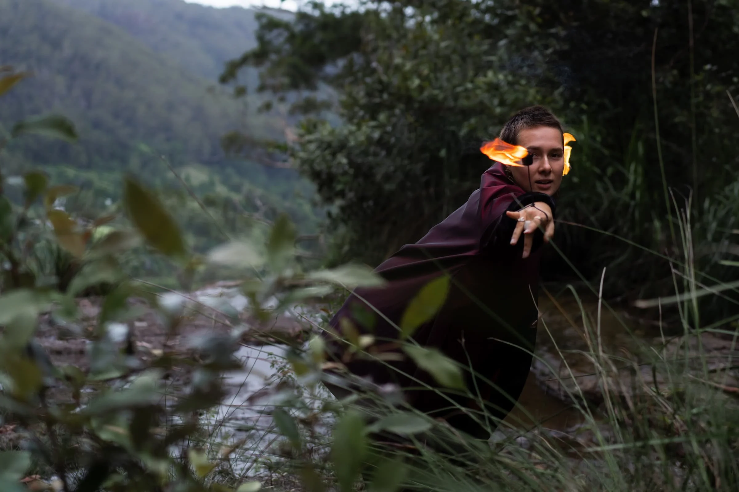A person with short dark hair, wearing a dark outfit, is in a swampy area surrounded by green foliage and trees, extending a hand forward with a flame on their ear.