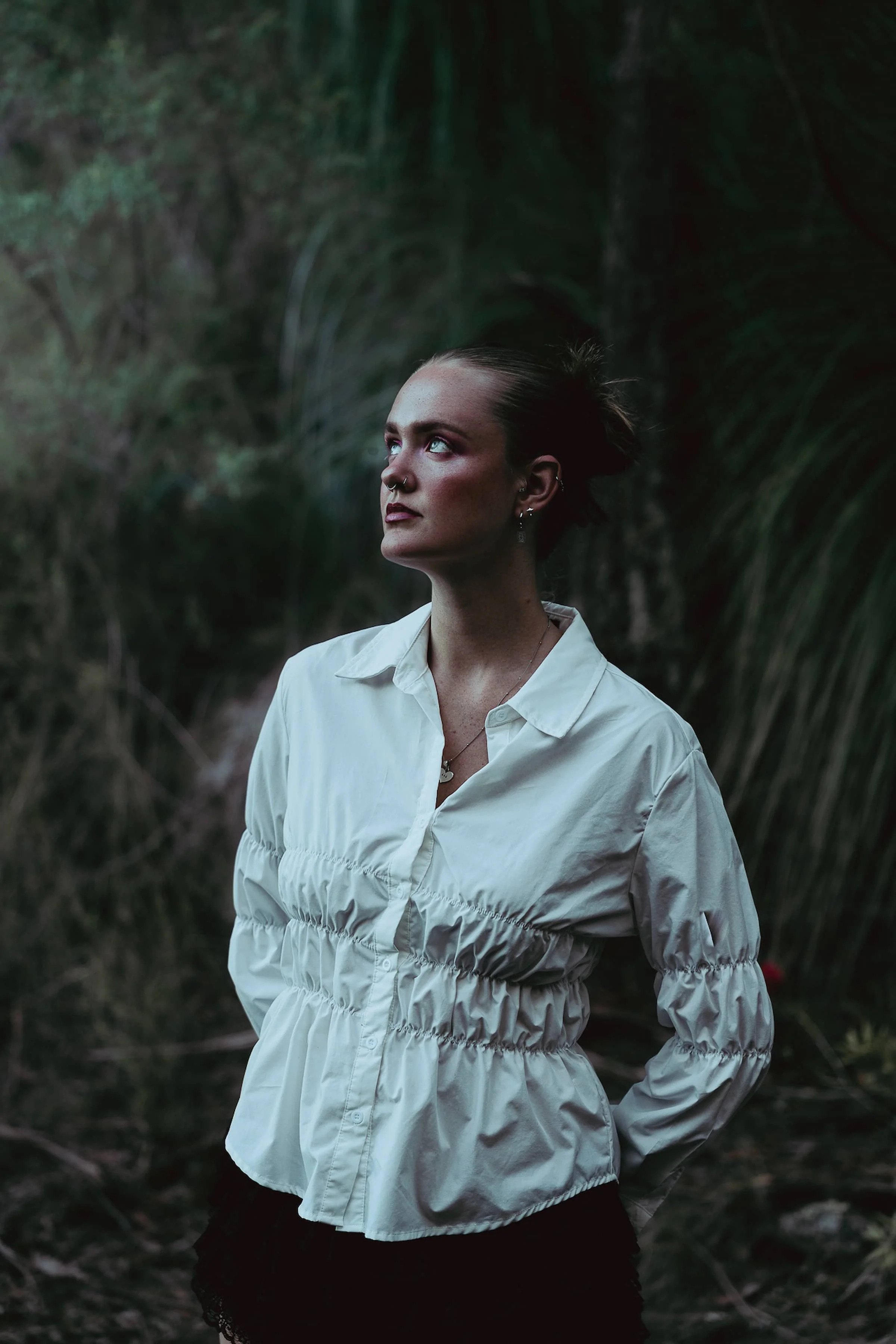 A woman with dark hair tied back, wearing a white gathered blouse, standing outdoors in a lush, green environment, looking to the side.