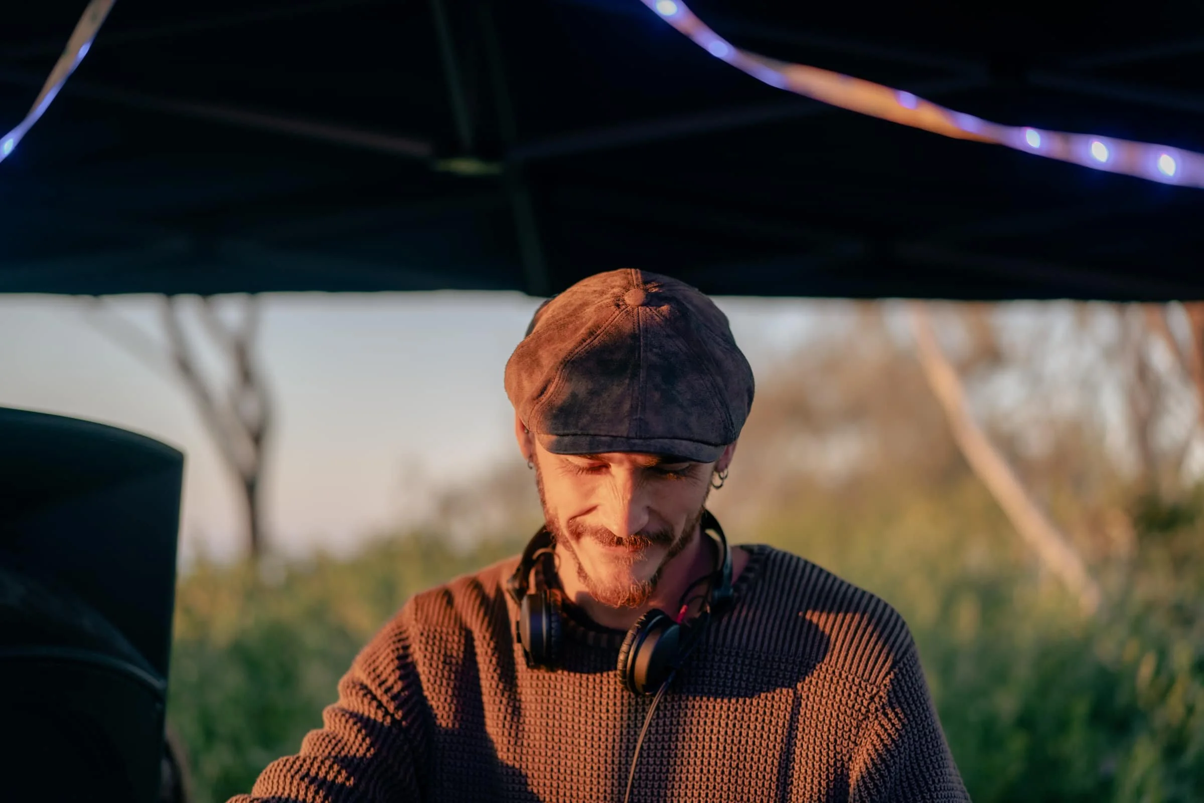 A young man with headphones around his neck, wearing a flat cap and a striped sweater, looking down while standing outdoors under a canopy or tent during sunset.